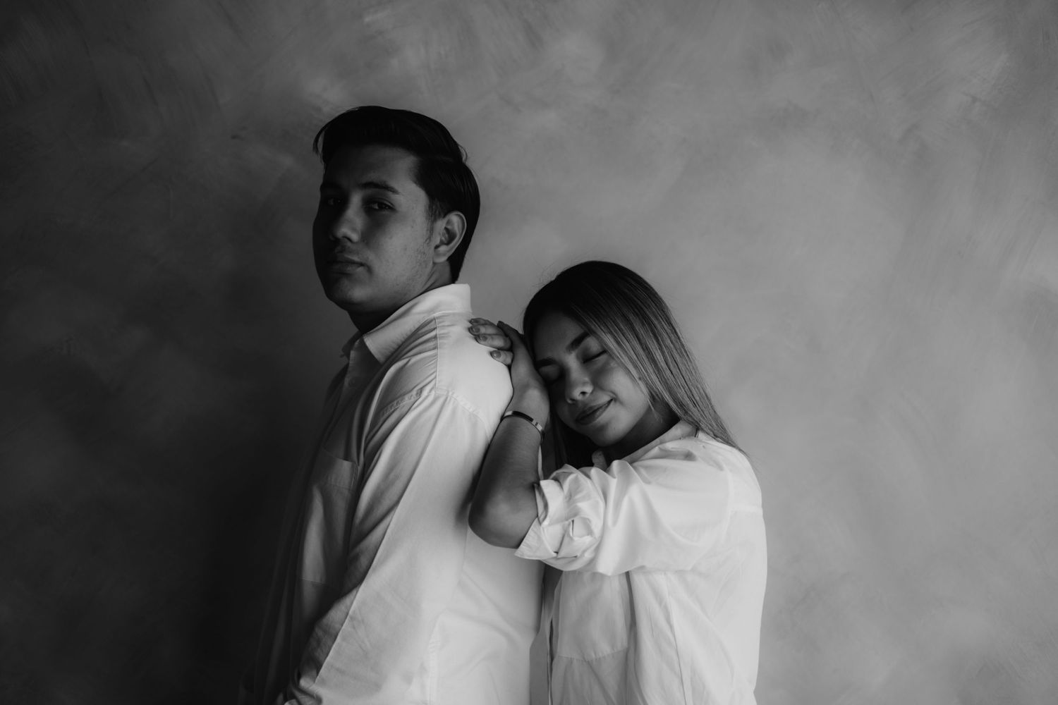 Black and white studio portrait of a couple, woman resting her head on mans shoulder from behind.