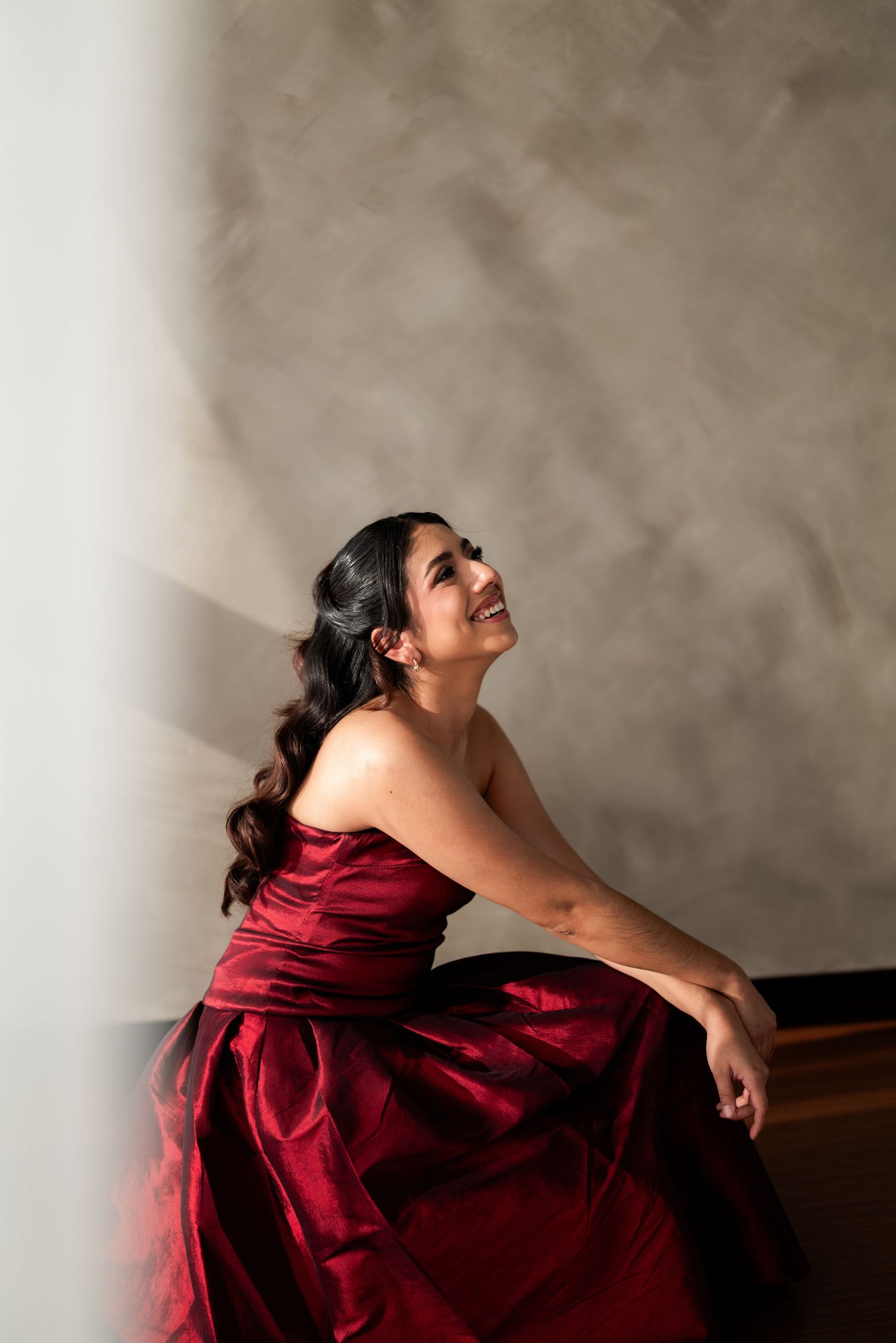 Radiant young woman in a stunning red strapless ball gown laughs joyfully in a sunlit studio.