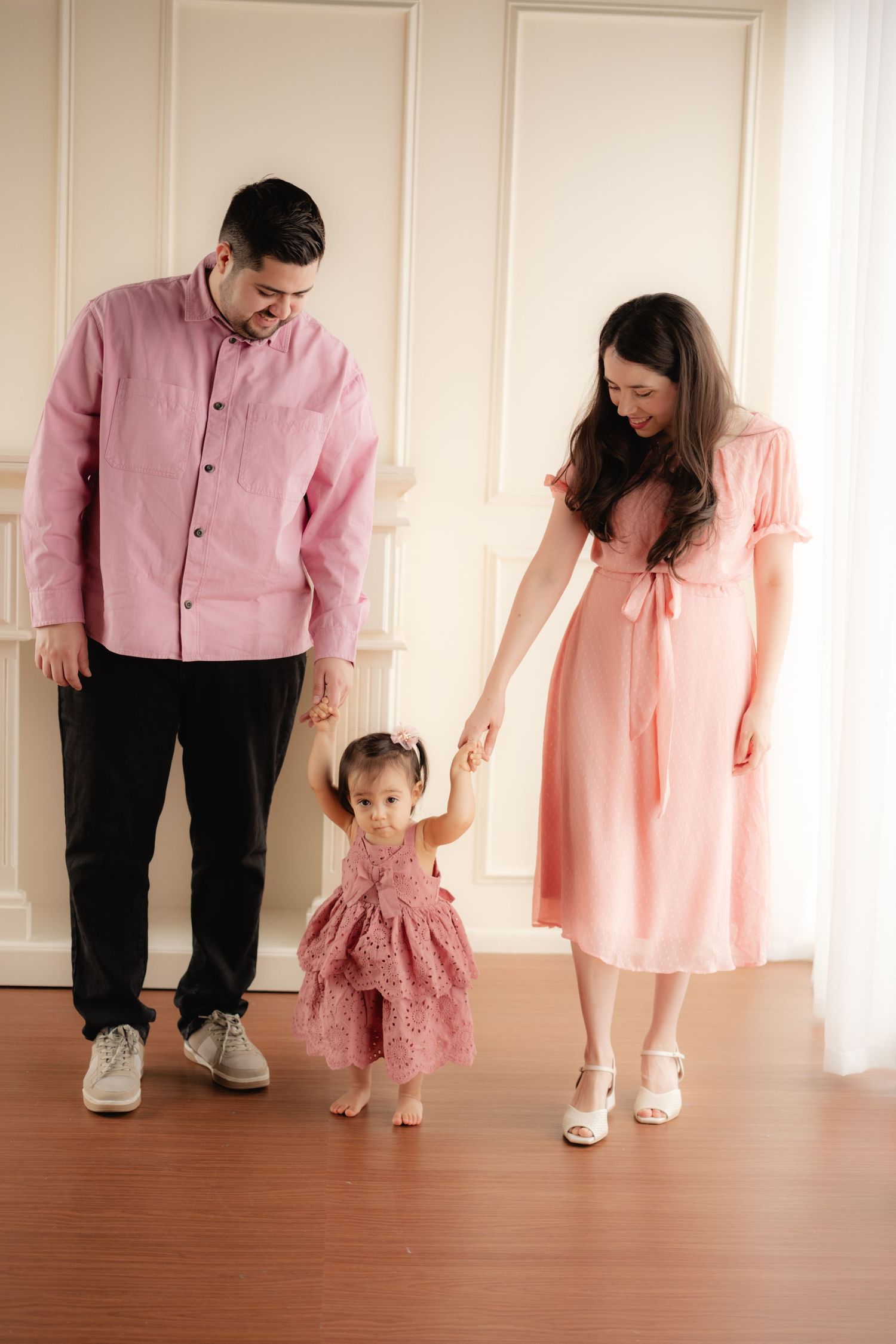 Parents in matching pink outfits help their toddler daughter take steps in a bright elegant room.