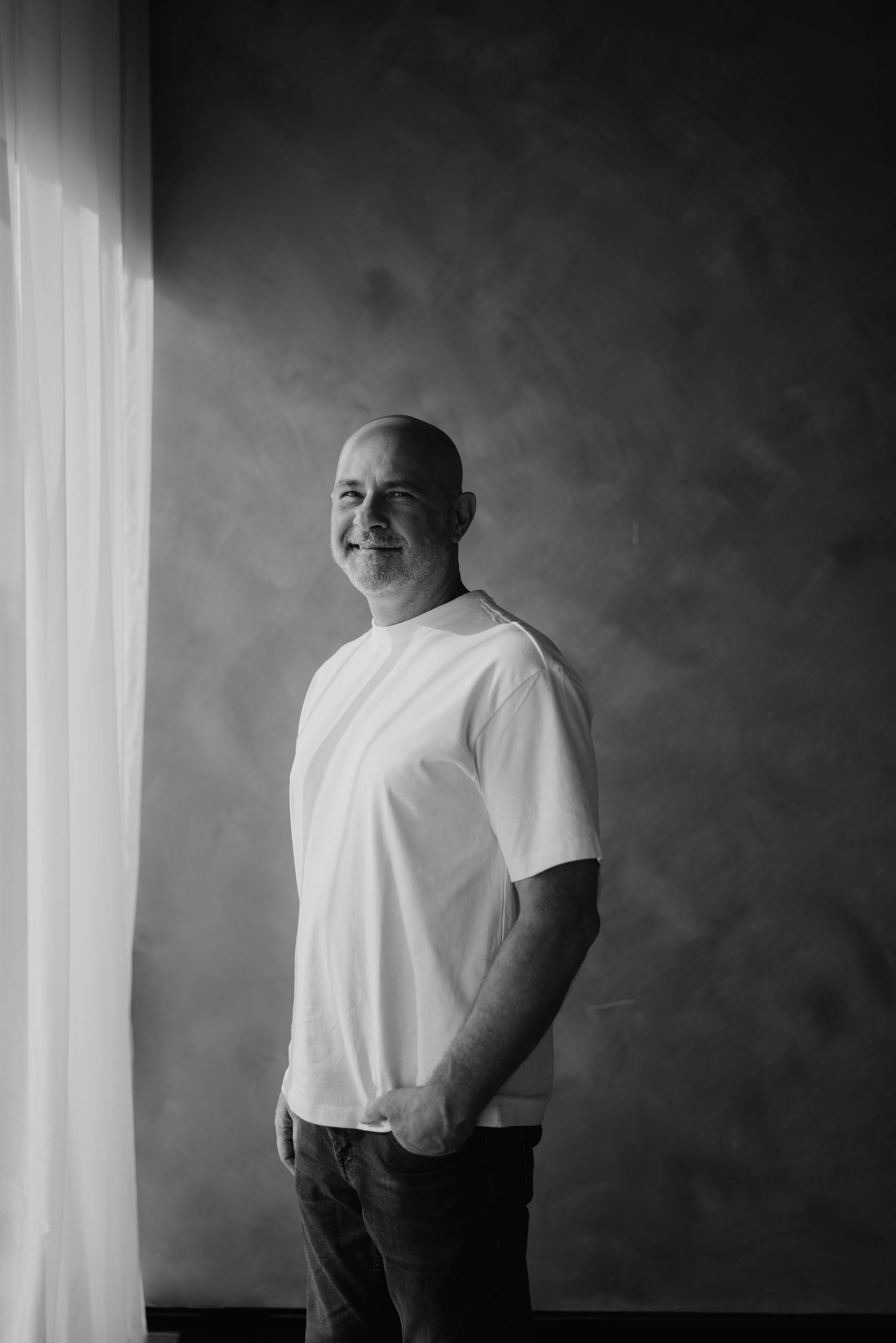 Black and white portrait of smiling bald man in white t-shirt standing by window.