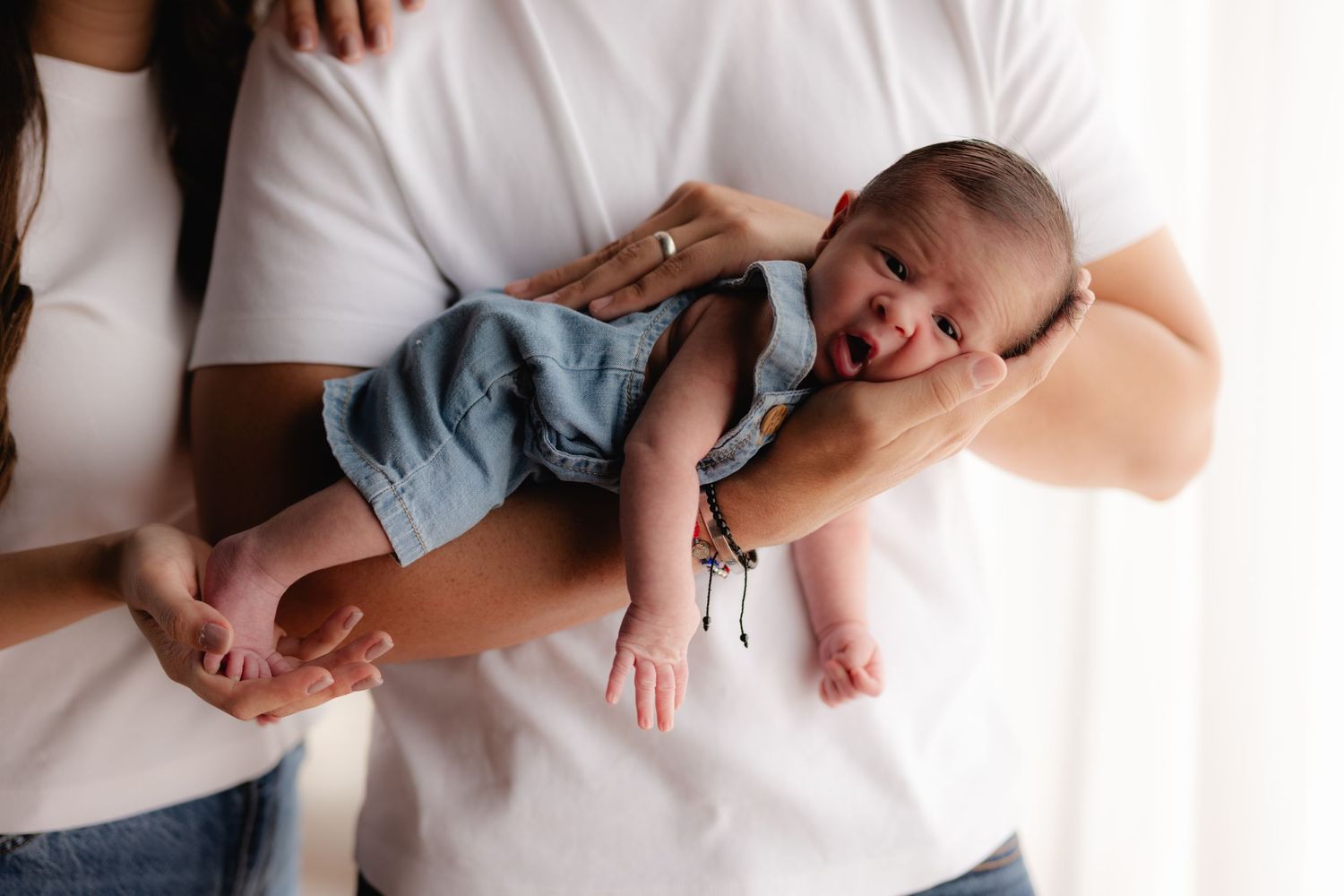 Parents in white shirts cradling alert newborn baby dressed in denim overalls near bright window.