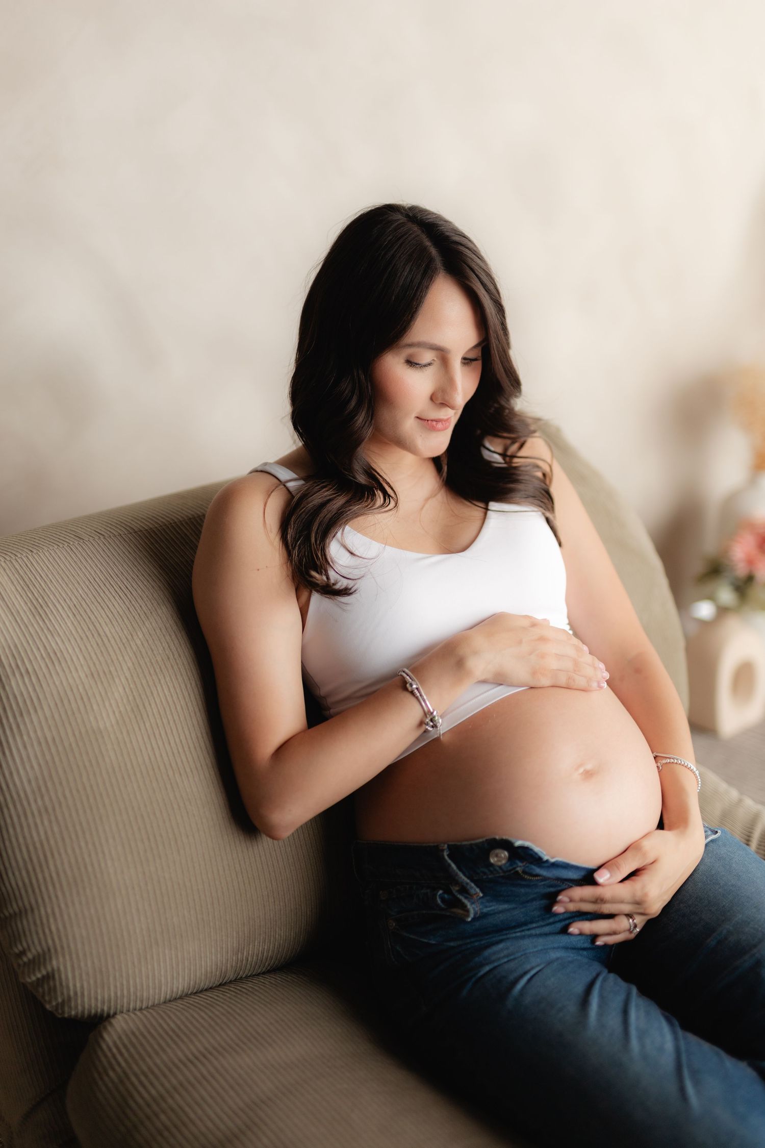 Pregnant woman in white crop top and jeans sitting on sofa, cradling her baby bump with both hands.