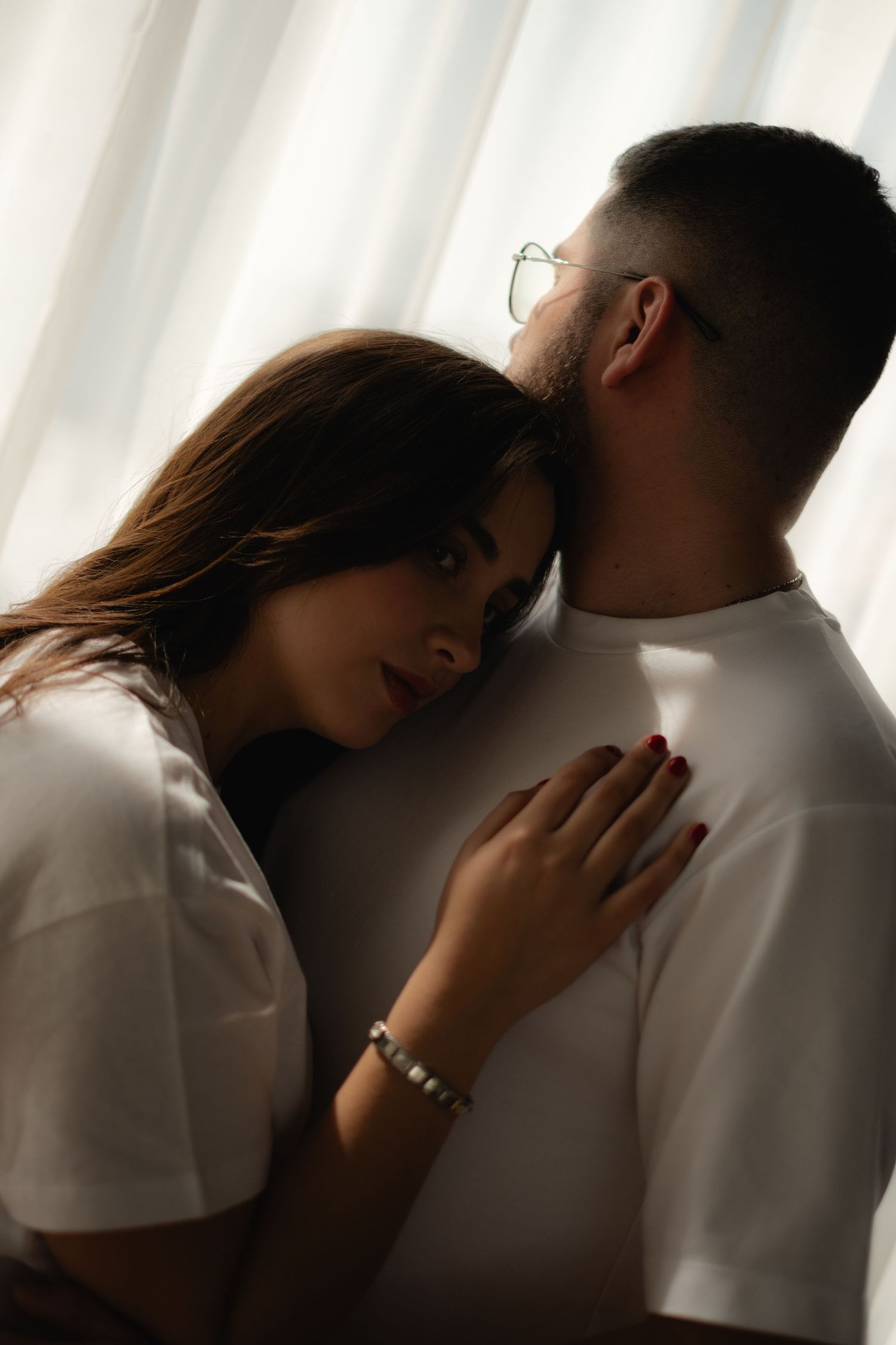 Intimate couple moment with woman resting head on mans chest while he kisses her forehead near white curtains.