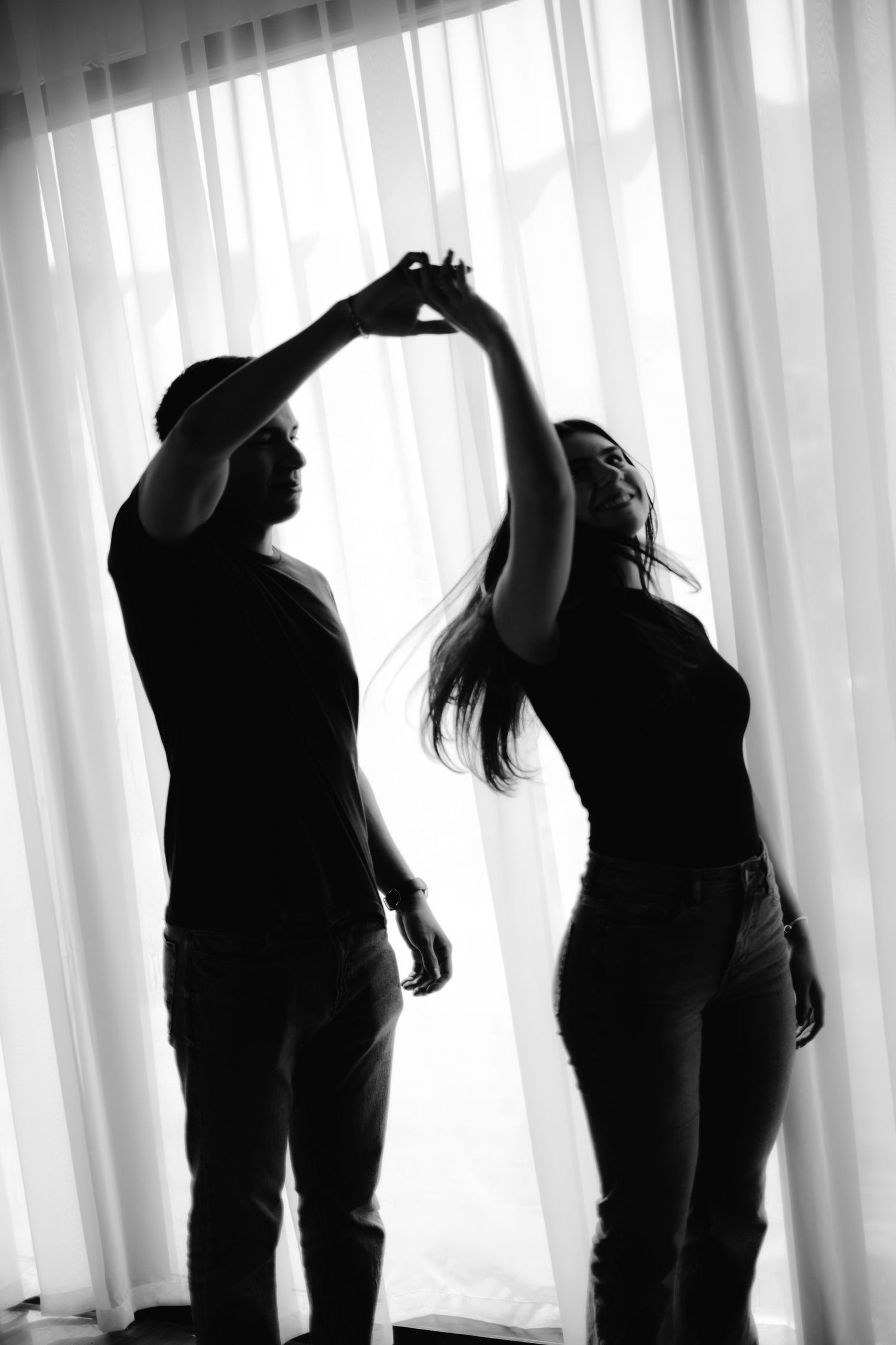 Silhouette of a couple dancing joyfully in front of bright backlit curtains in black and white.