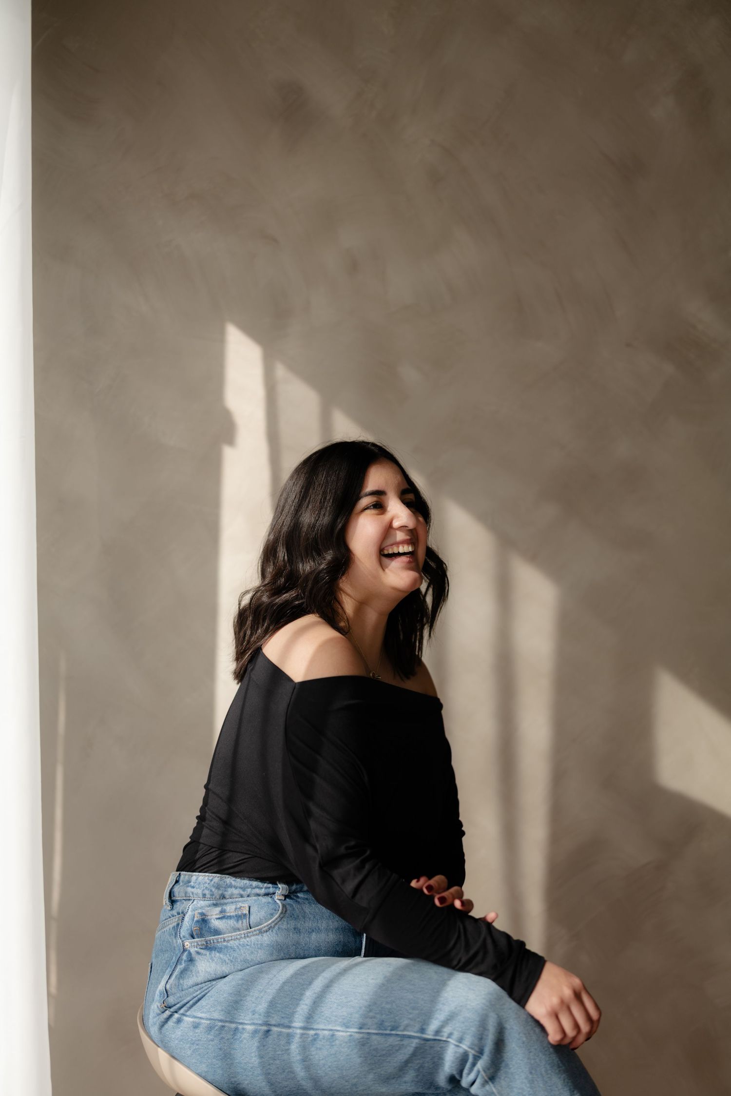Joyful young woman in black off-shoulder top laughs while sitting in warm natural sunlight indoors.