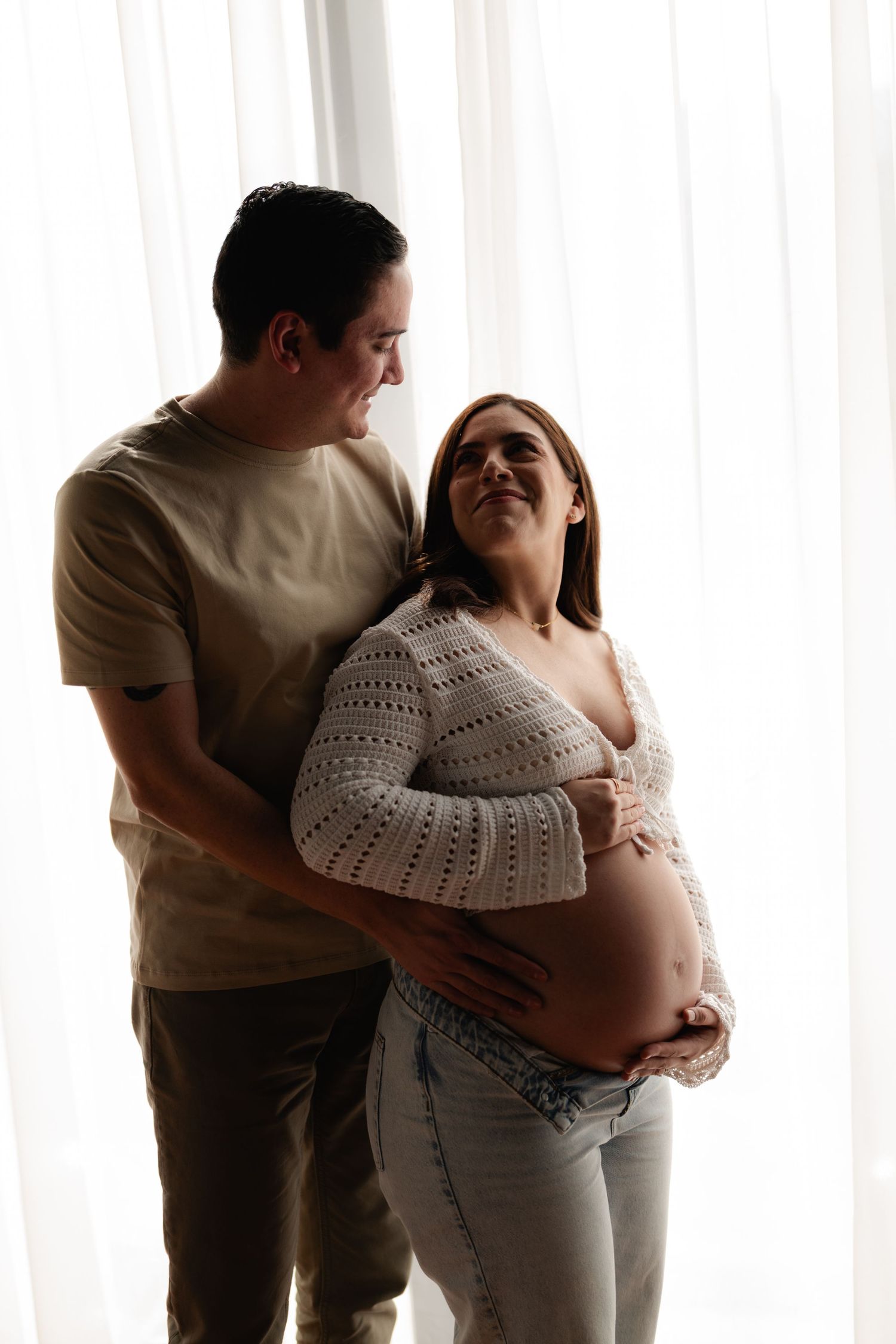 Man embraces smiling pregnant woman from behind near sheer white curtains in a maternity portrait.