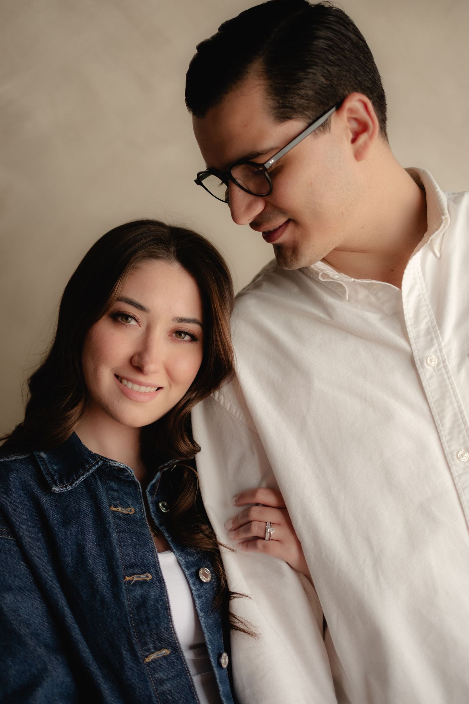 Woman in denim jacket smiles at camera while man in glasses and white shirt looks down at her tenderly.