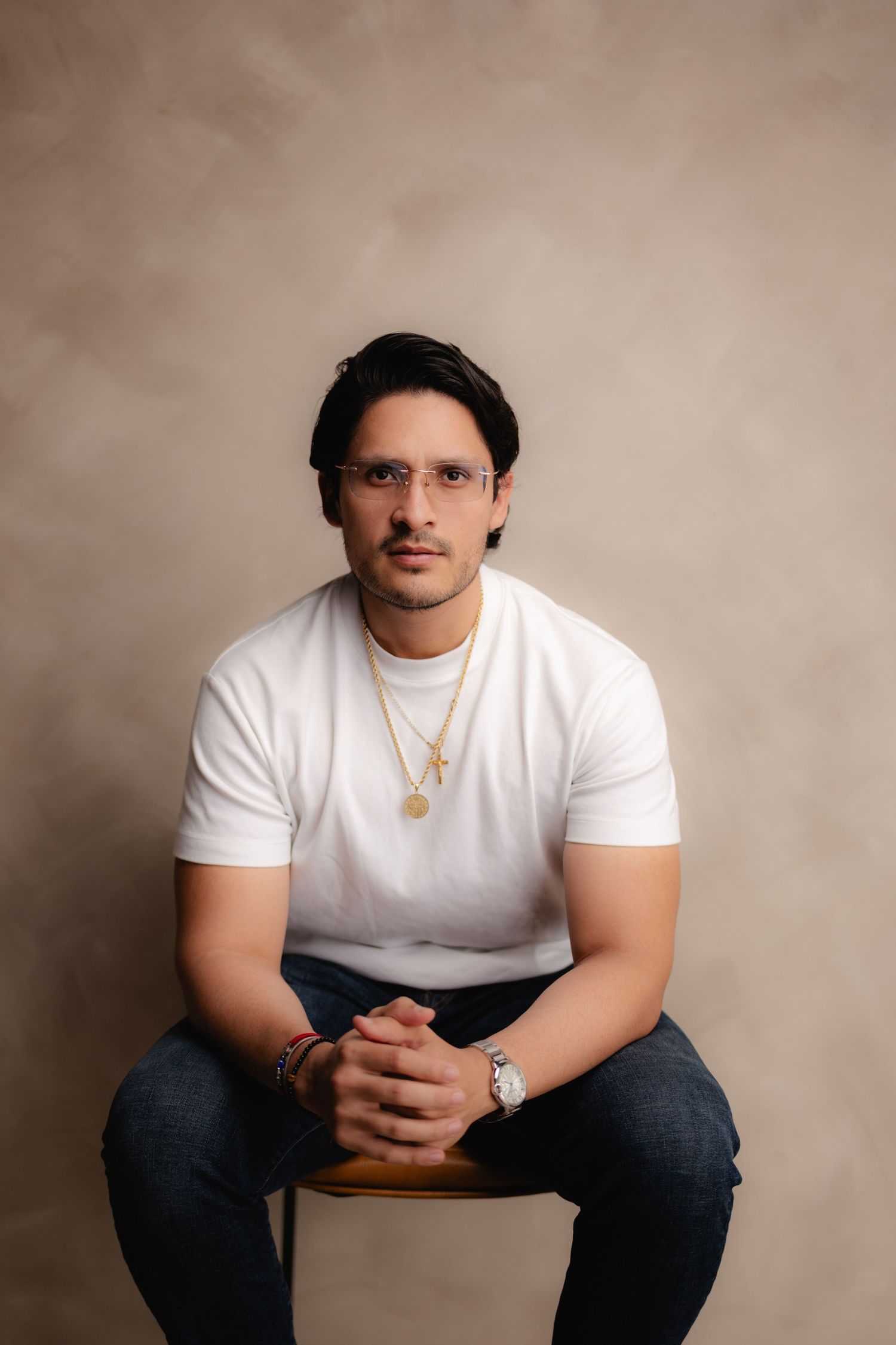 Man in white t-shirt with gold necklace sitting with clasped hands on a stool against beige backdrop.