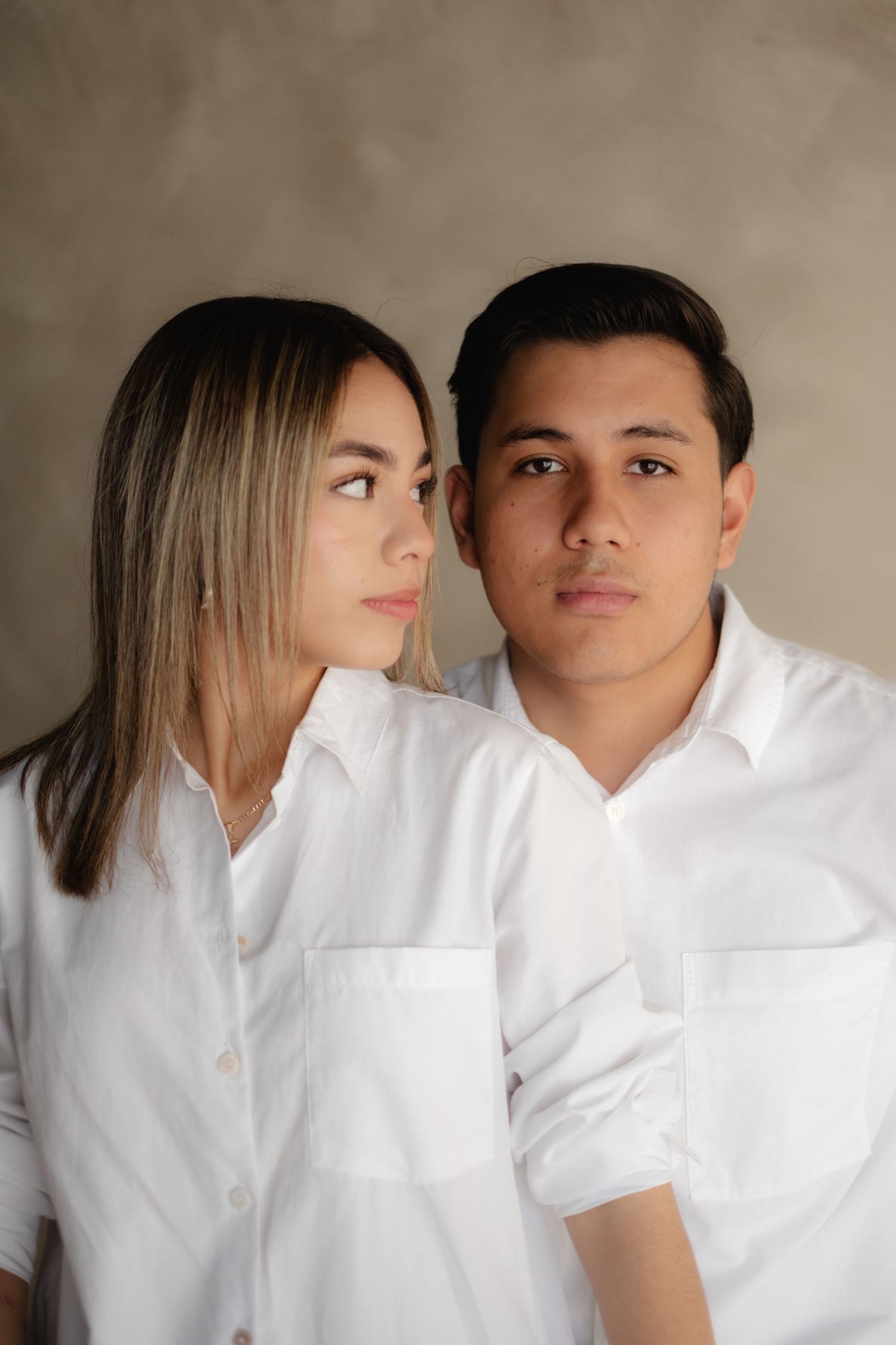 A young couple in matching white button-up shirts pose together against a gray background.