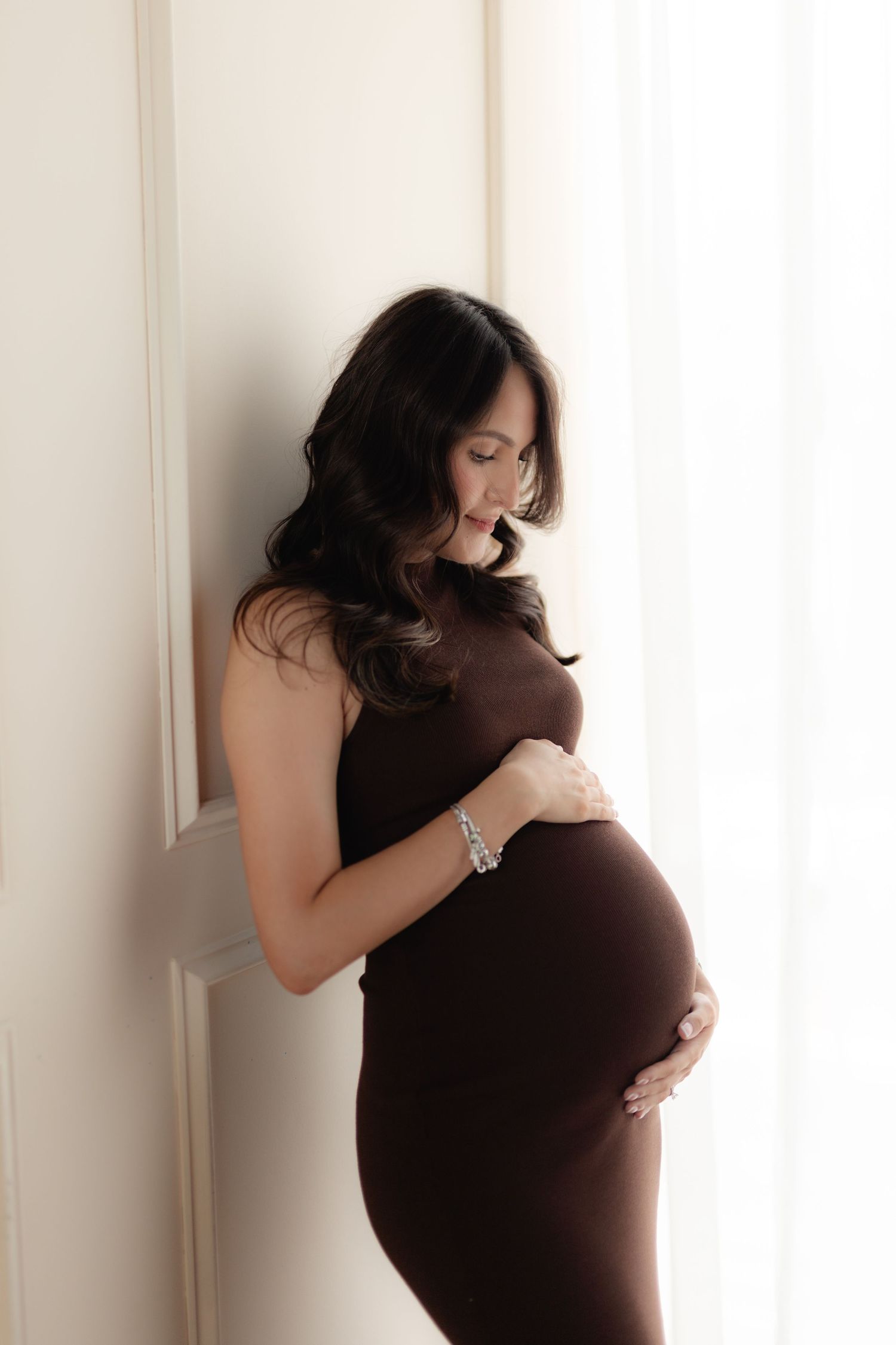 Pregnant woman in brown dress cradling baby bump, leaning against white wall in soft natural light.