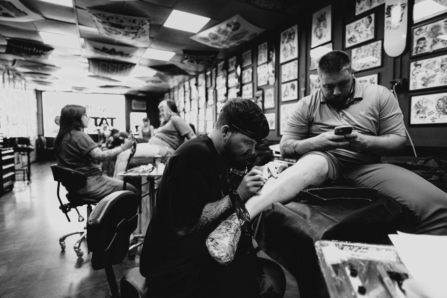 Black and white photo of a tattoo artist tattooing client's leg, with more tattoo artist visible in the background