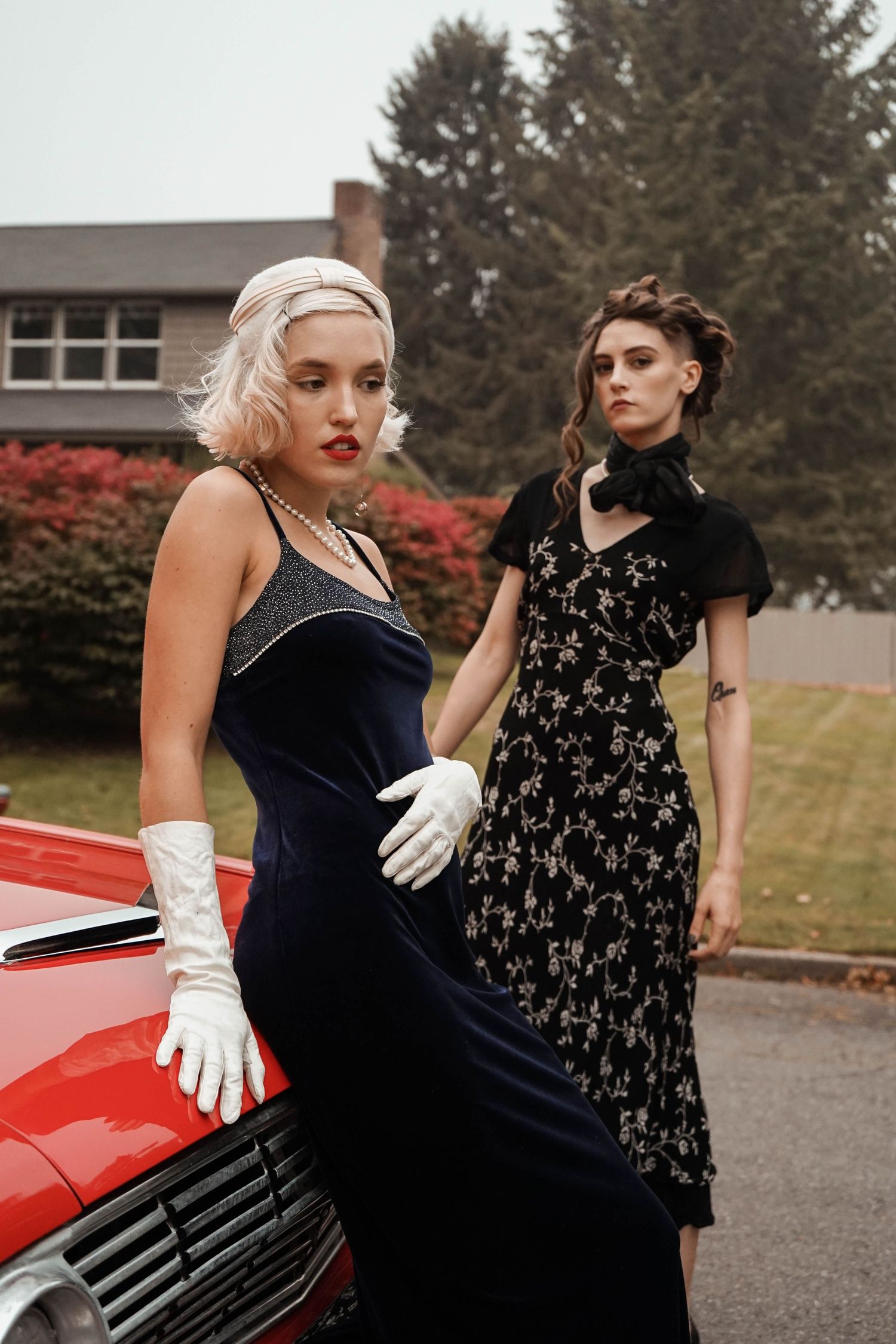 Creative portrait of two women in timeless dresses posing on a red car in a neighborhood