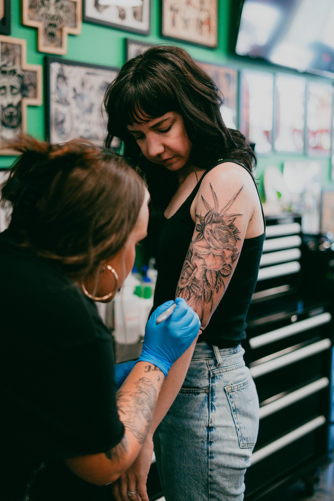 A tattoo artist drawing on a girls arm while the girl looks and watches the tattoo artist