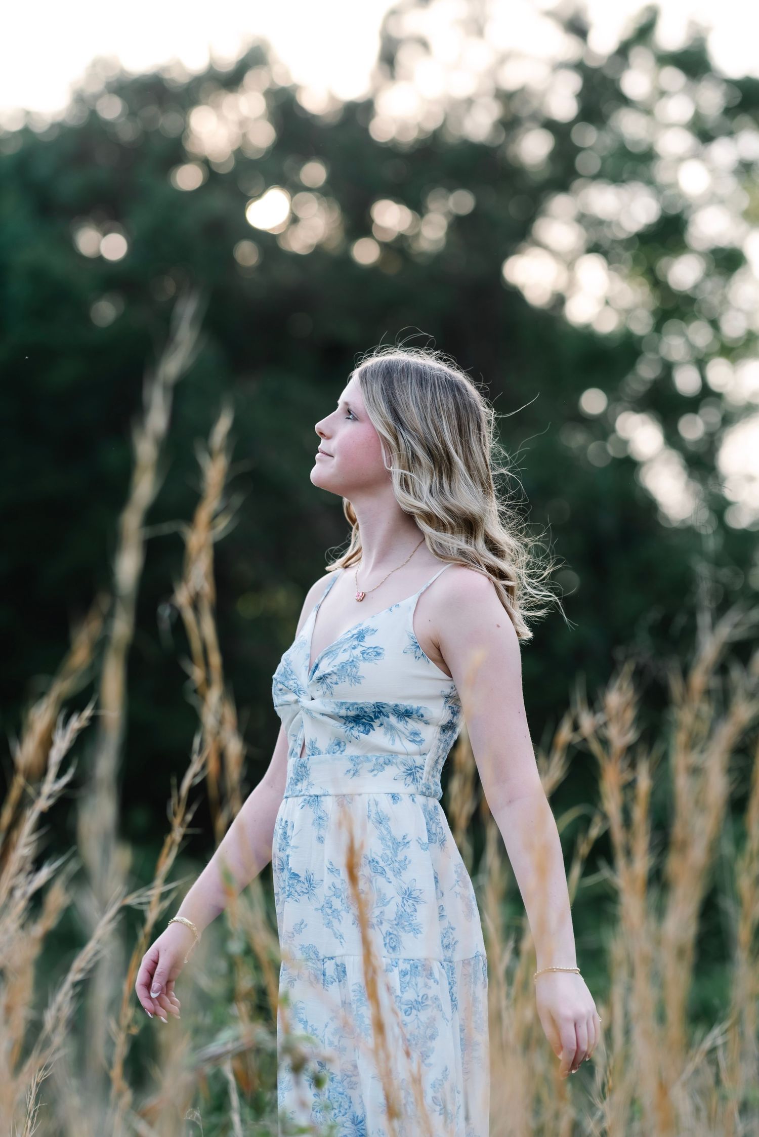 Young woman in floral maxi dress standing in golden tall grass field with trees in background.
