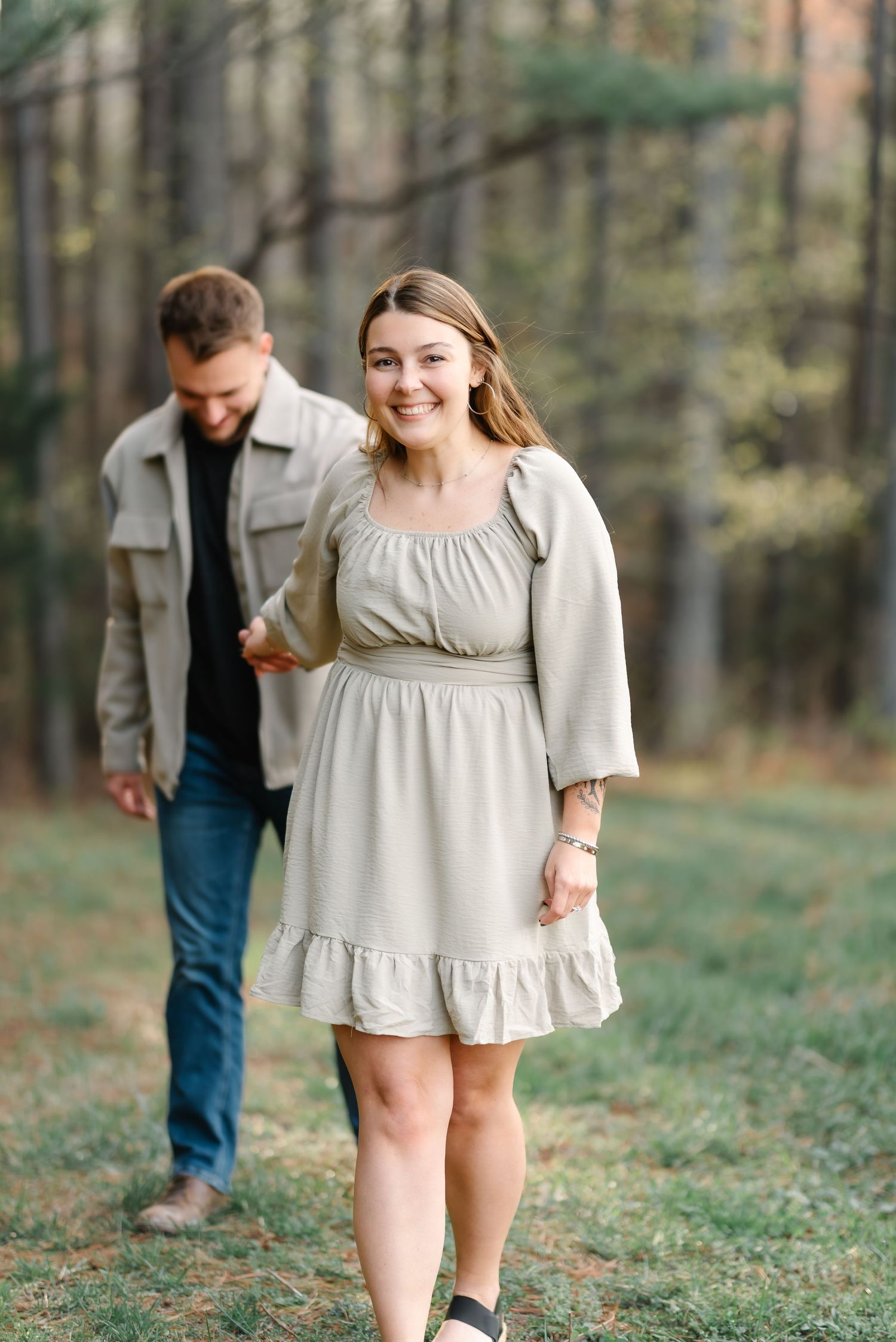 A smiling woman in a beige dress holds hands with a man in a jacket while walking through a forest path.