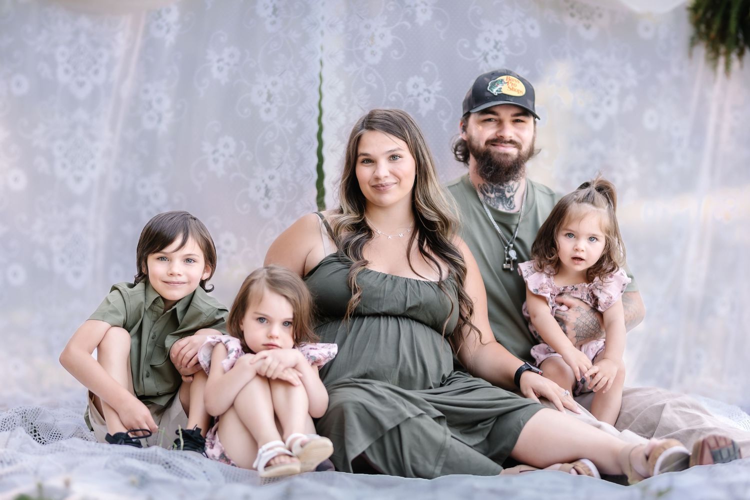 A family of five poses together in matching olive green outfits against a soft floral backdrop for a maternity photo session.