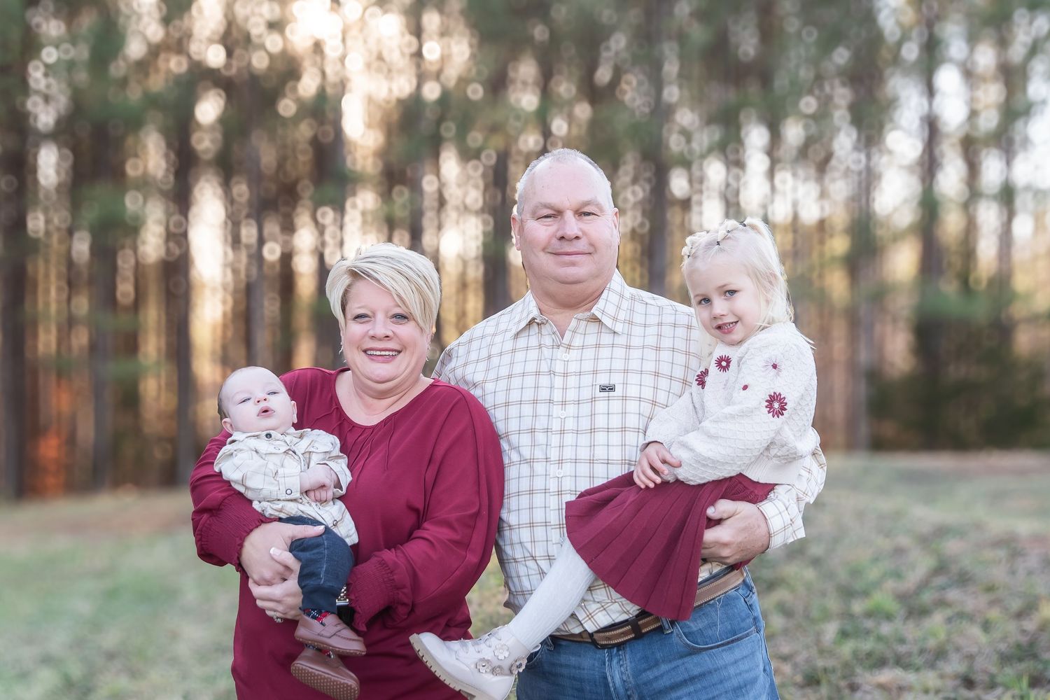 Family of four posing outdoors among tall pine trees, grandparents holding two young children in coordinating red and white outfits.