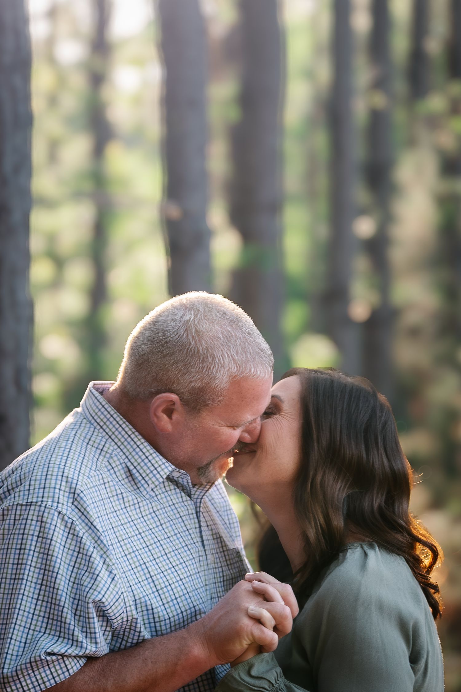 Couple sharing a romantic kiss in a sunlit forest, man in plaid shirt and woman in green top holding hands.