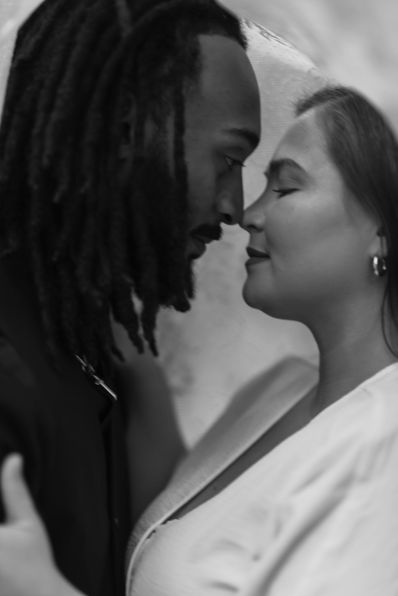 Black and white photo of a romantic couple sharing an intimate moment, man with dreadlocks and woman in white nearly kissing.
