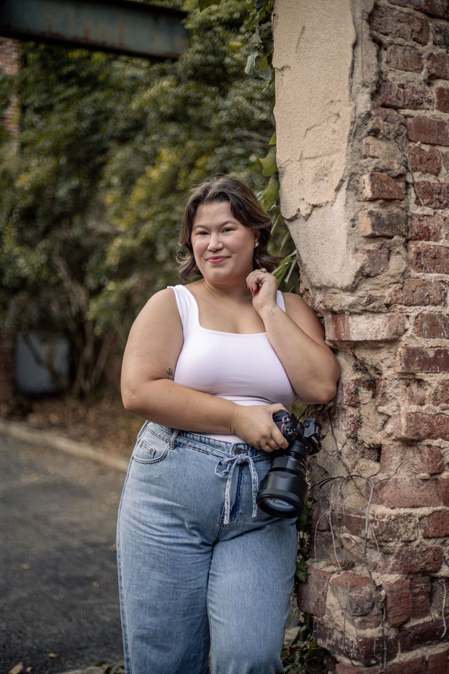 Young woman in white crop top and jeans holding a camera, leaning against a brick wall outdoors.