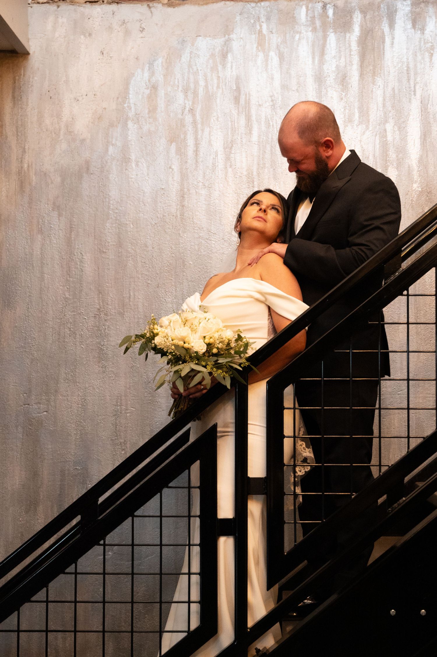 Bride and groom share a romantic moment on a staircase, she holds white bouquet while gazing up at him in black tuxedo.