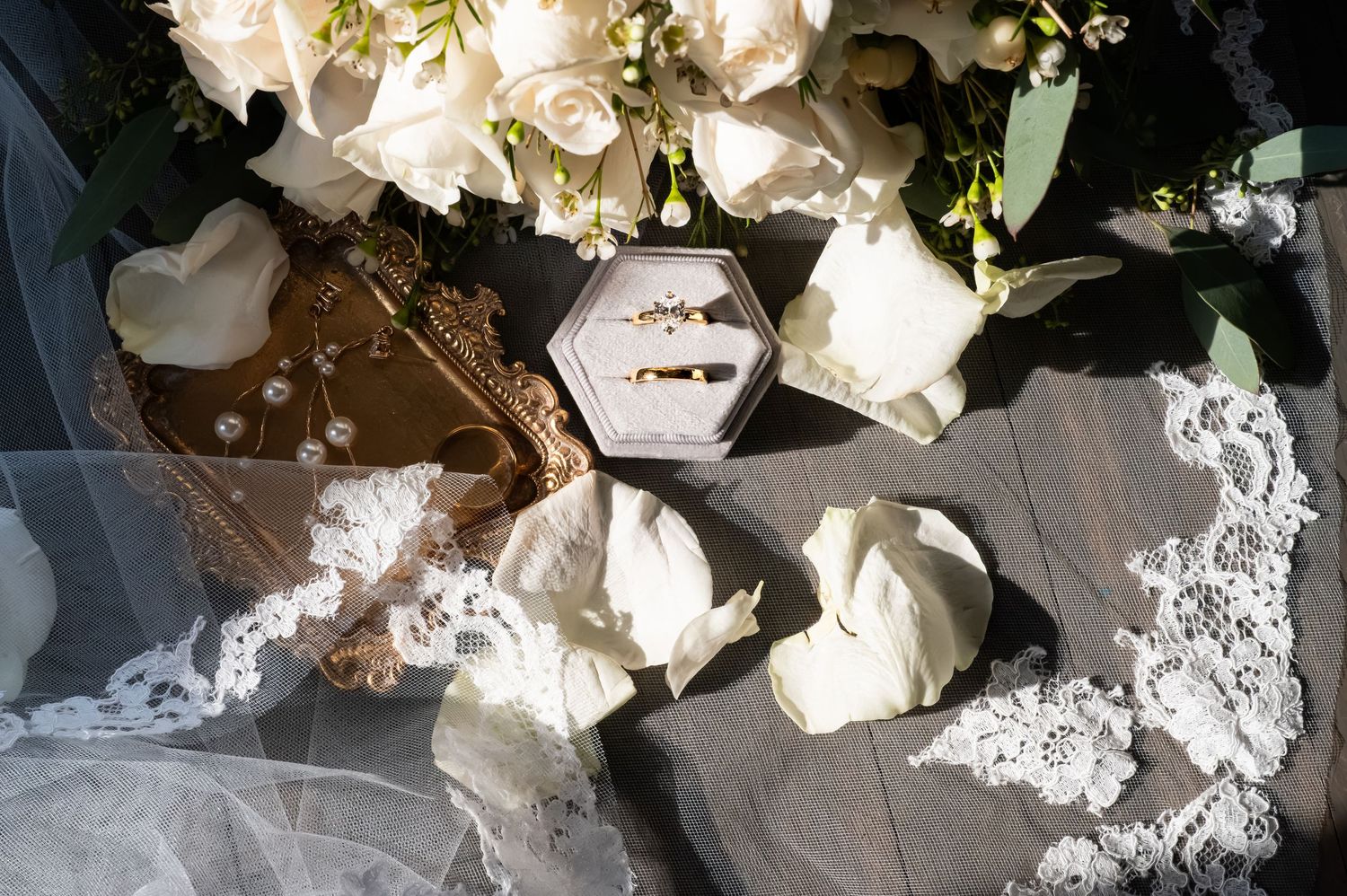Two gold wedding rings in a hexagonal velvet box surrounded by white roses, lace veil, and rose petals.