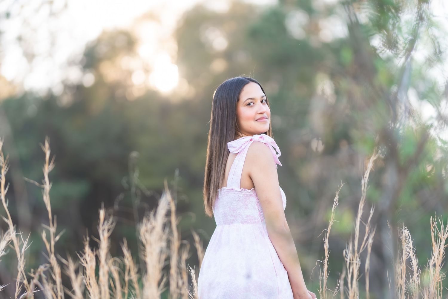 Young woman in white dress standing in golden field with soft bokeh trees background in warm sunset light.