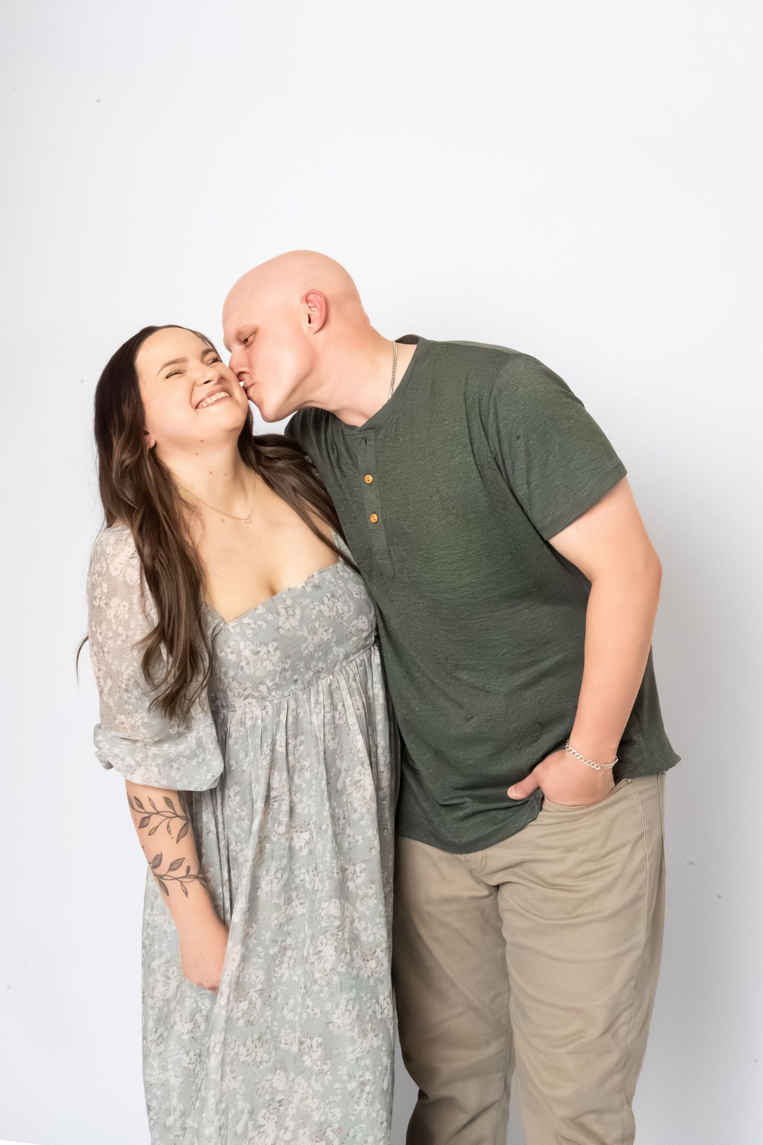 A bald man in a green shirt kisses a smiling woman with a tattoo in a floral dress against a white background.