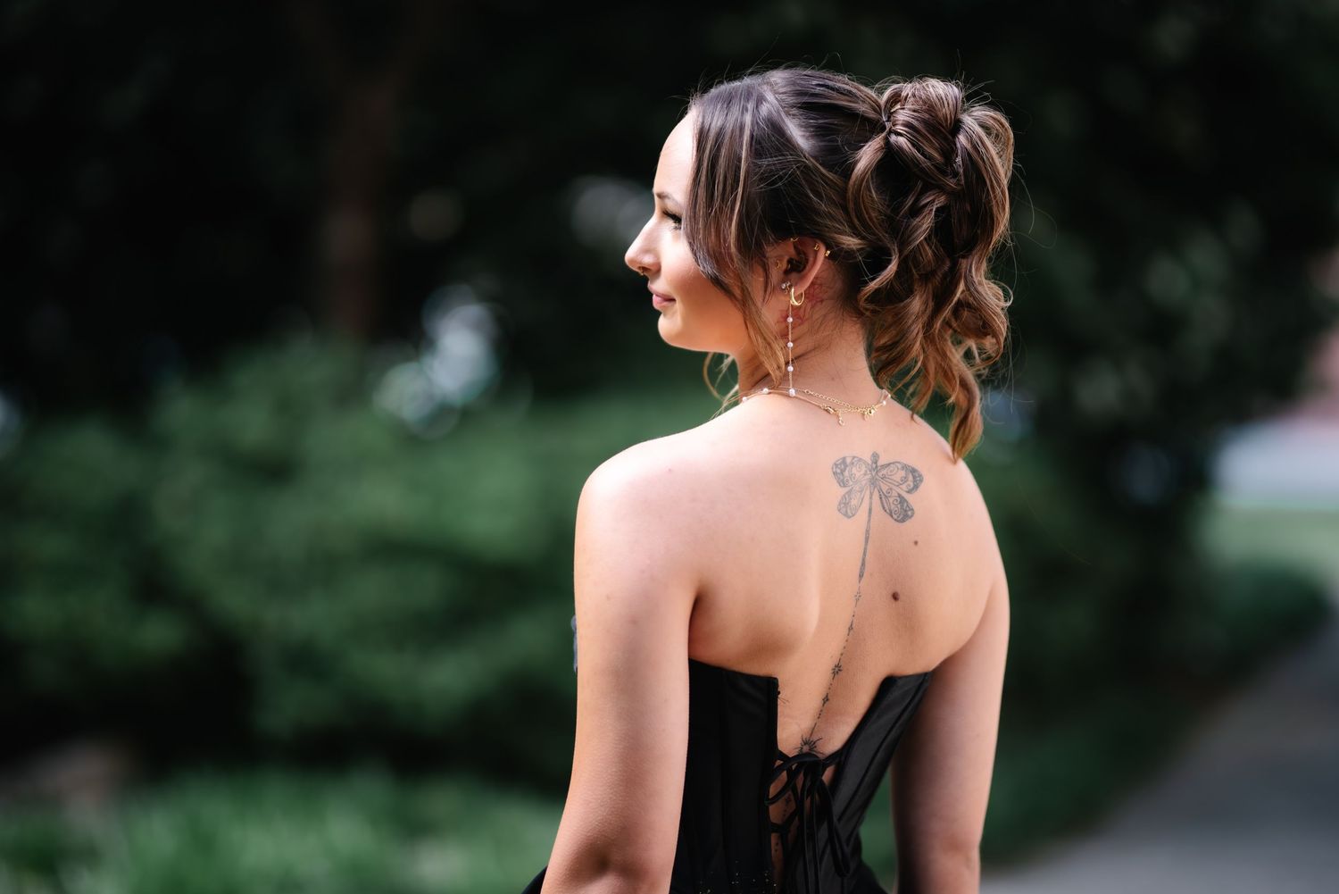 Young woman in black backless dress with dragonfly tattoo on upper back, hair up, standing outdoors.