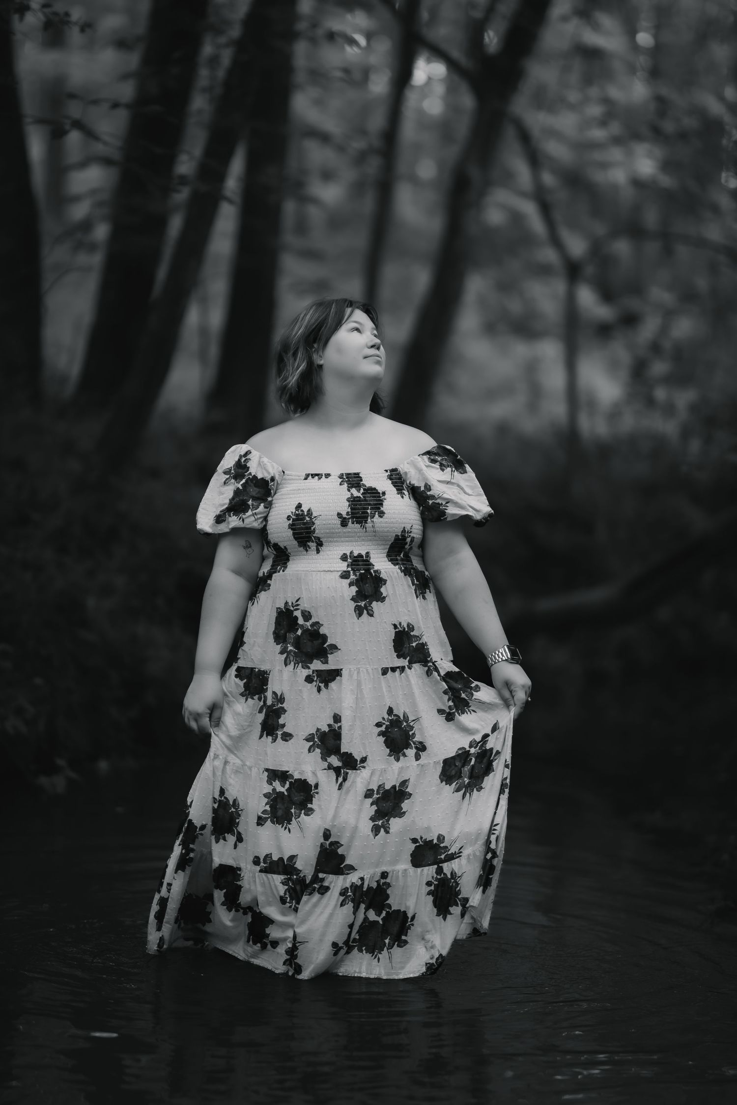 Black and white portrait of a woman in a floral off-shoulder dress standing in a forest stream.