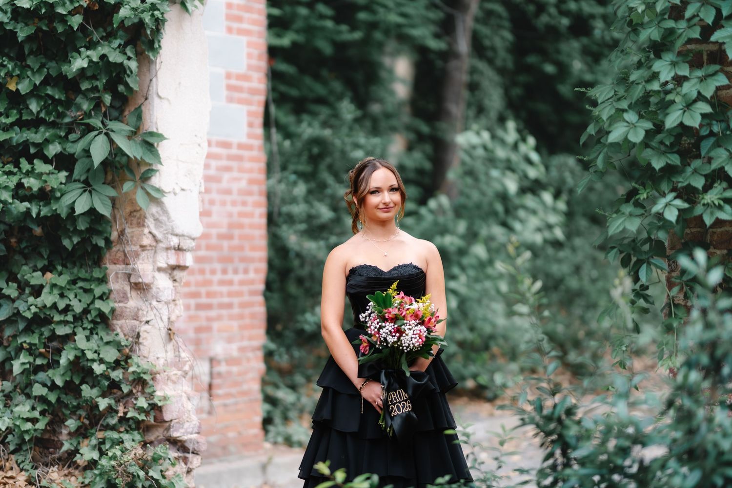 Woman in black strapless dress holding colorful bouquet standing among lush green ivy near brick wall.