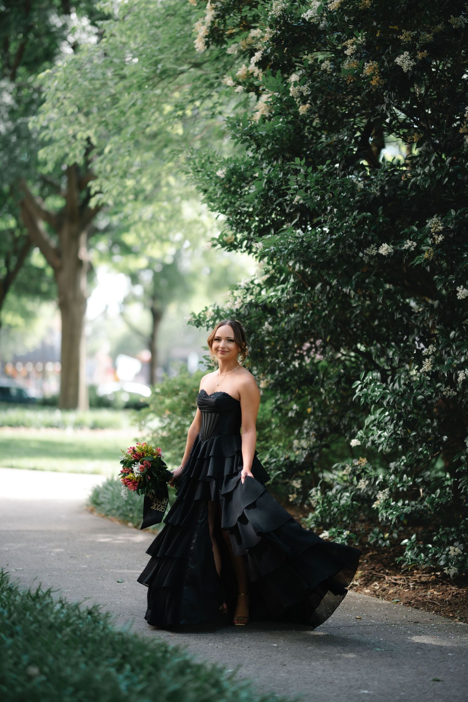 Woman in black strapless ruffled gown holding bouquet on tree-lined path.