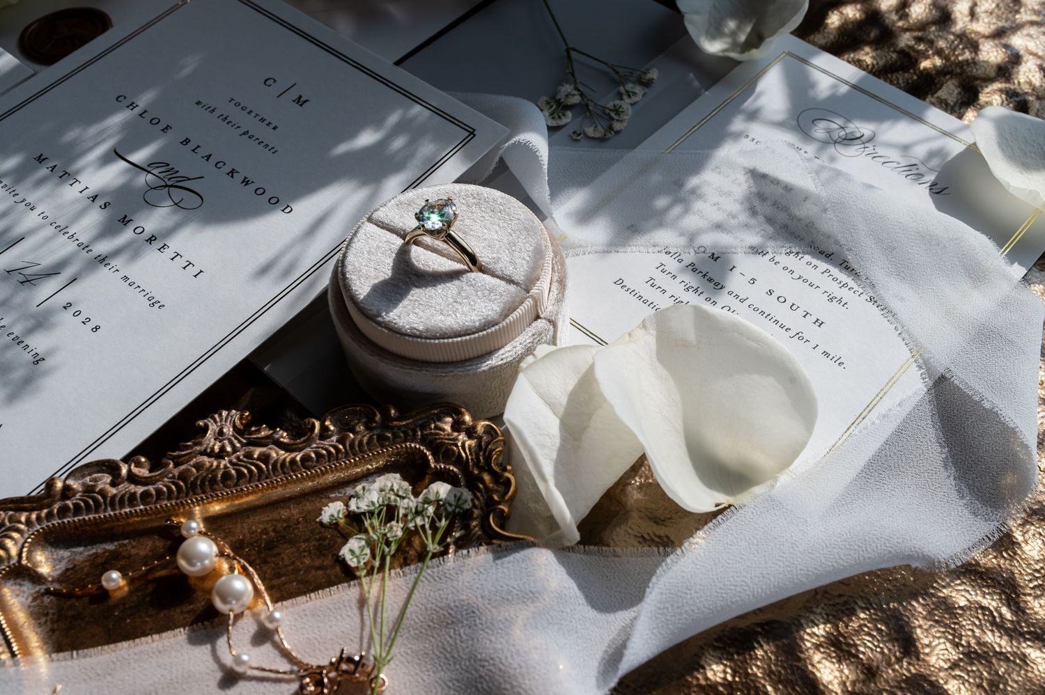 Elegant wedding flat lay with ring box, jewelry, florals, and stationery on white linen and gold tray.