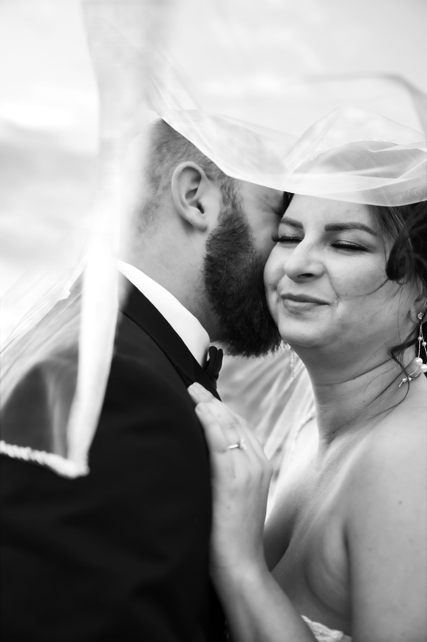 Black and white wedding photo of groom kissing bride on cheek under veil, bride smiling softly.