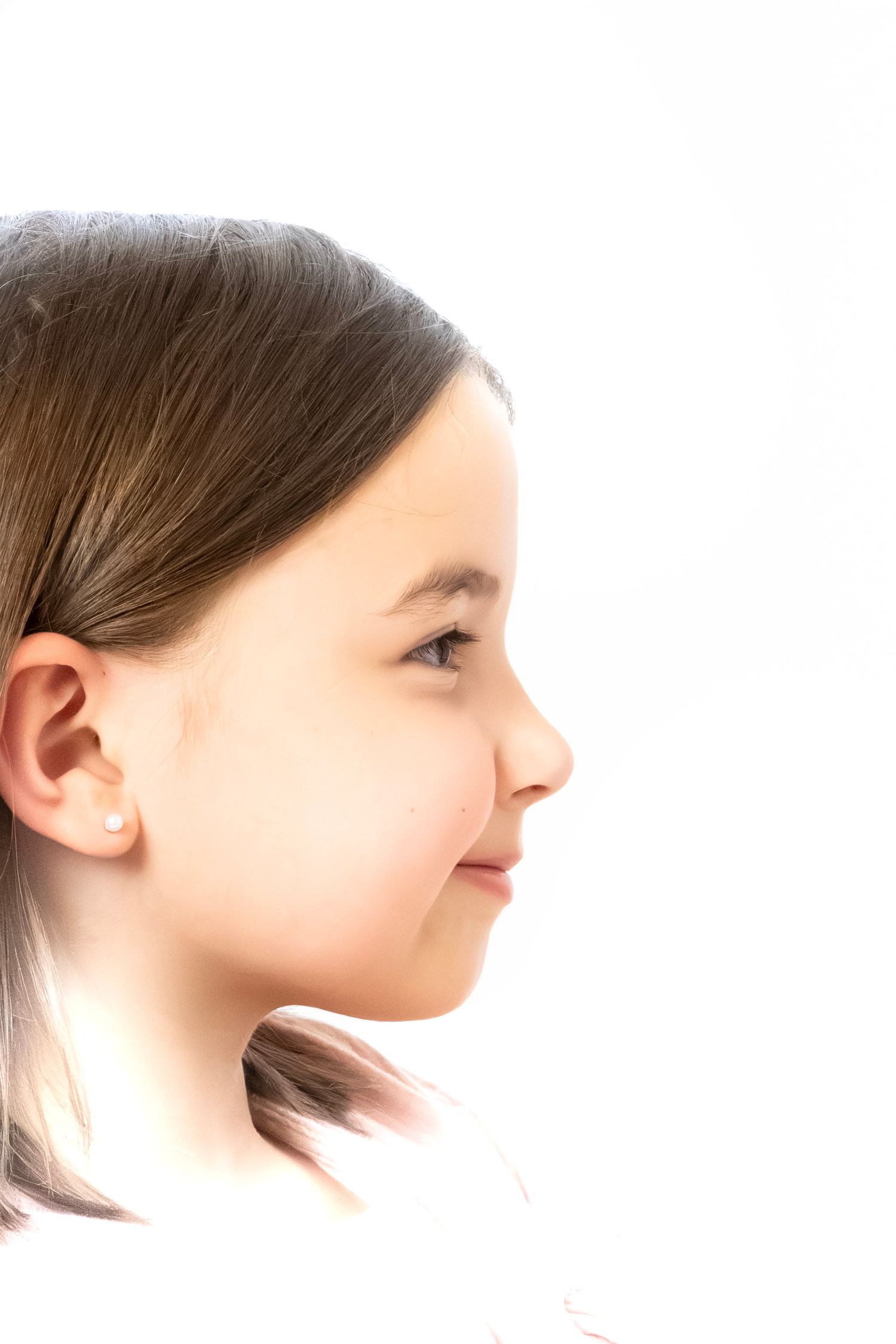 Profile view of a young girl with brown hair and stud earring, smiling softly against a white background.
