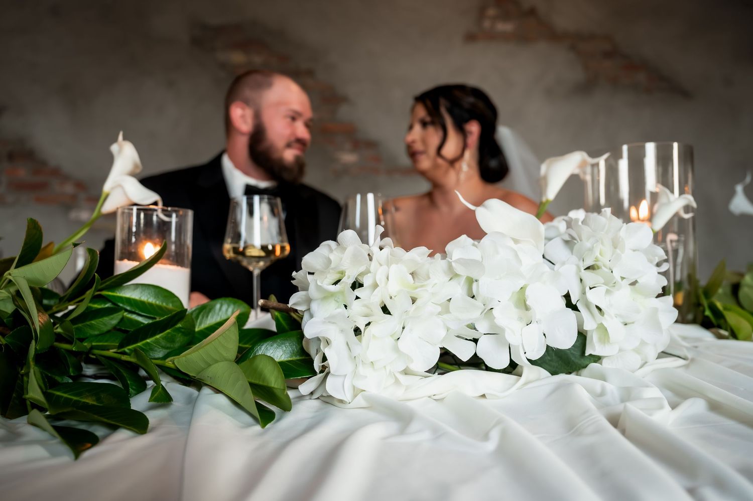 Bride and groom seated at wedding reception table with white floral bouquet and greenery garland in foreground.