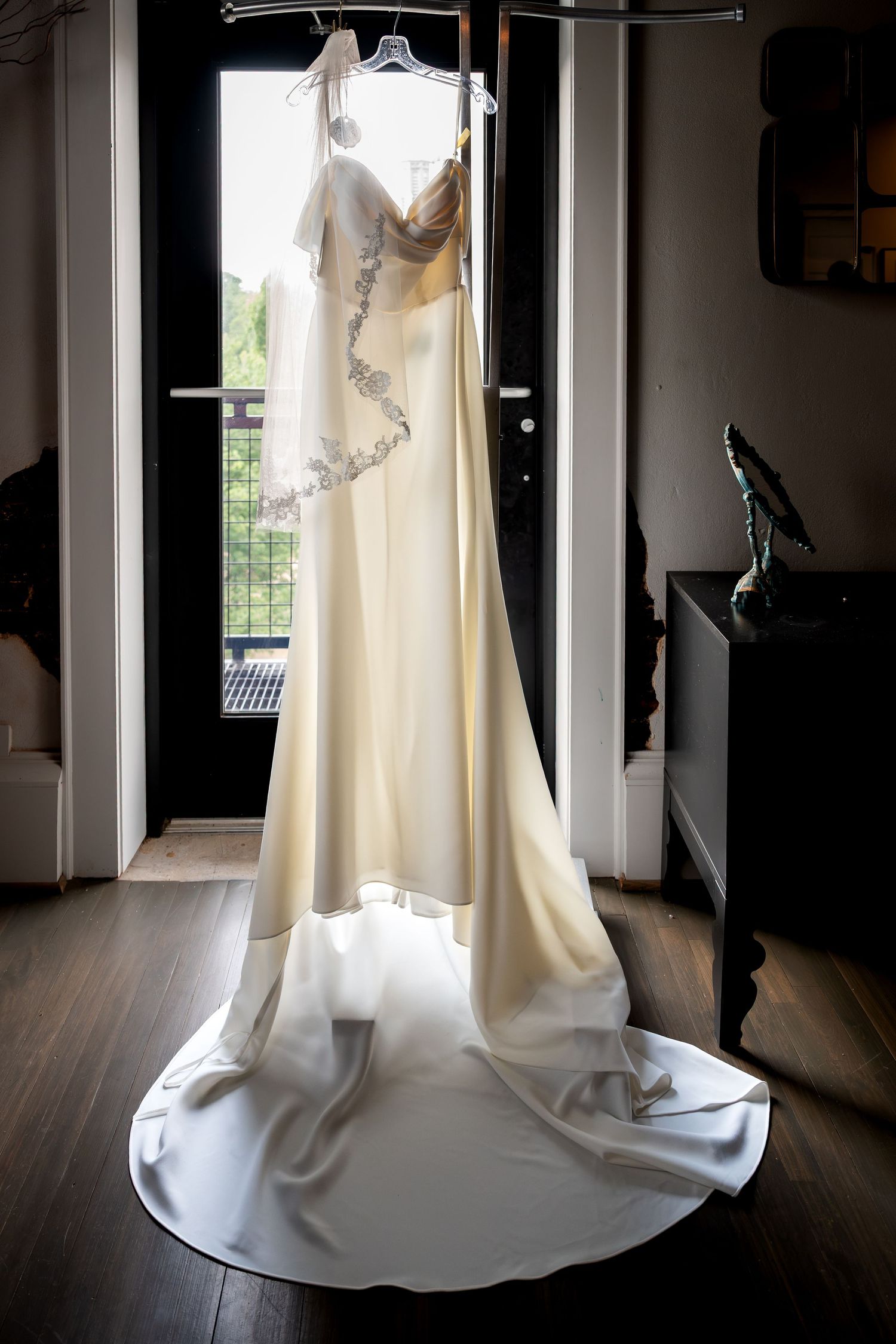 Elegant white wedding gown with floral lace detail hanging by a sunlit doorway, showcasing a long train on hardwood floors.