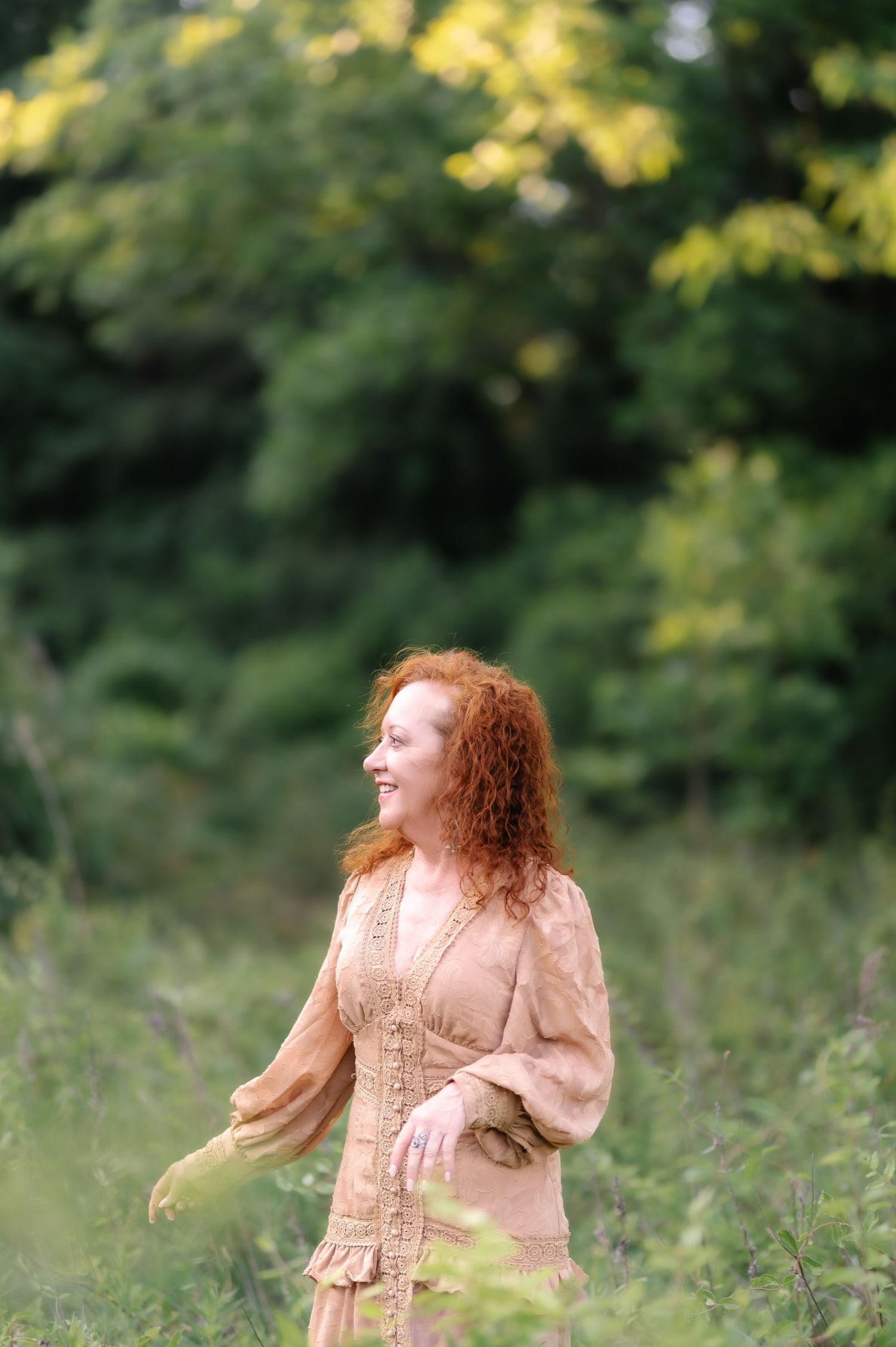 A smiling redhead woman in a beige boho dress stands outdoors surrounded by lush green trees and wildflowers.