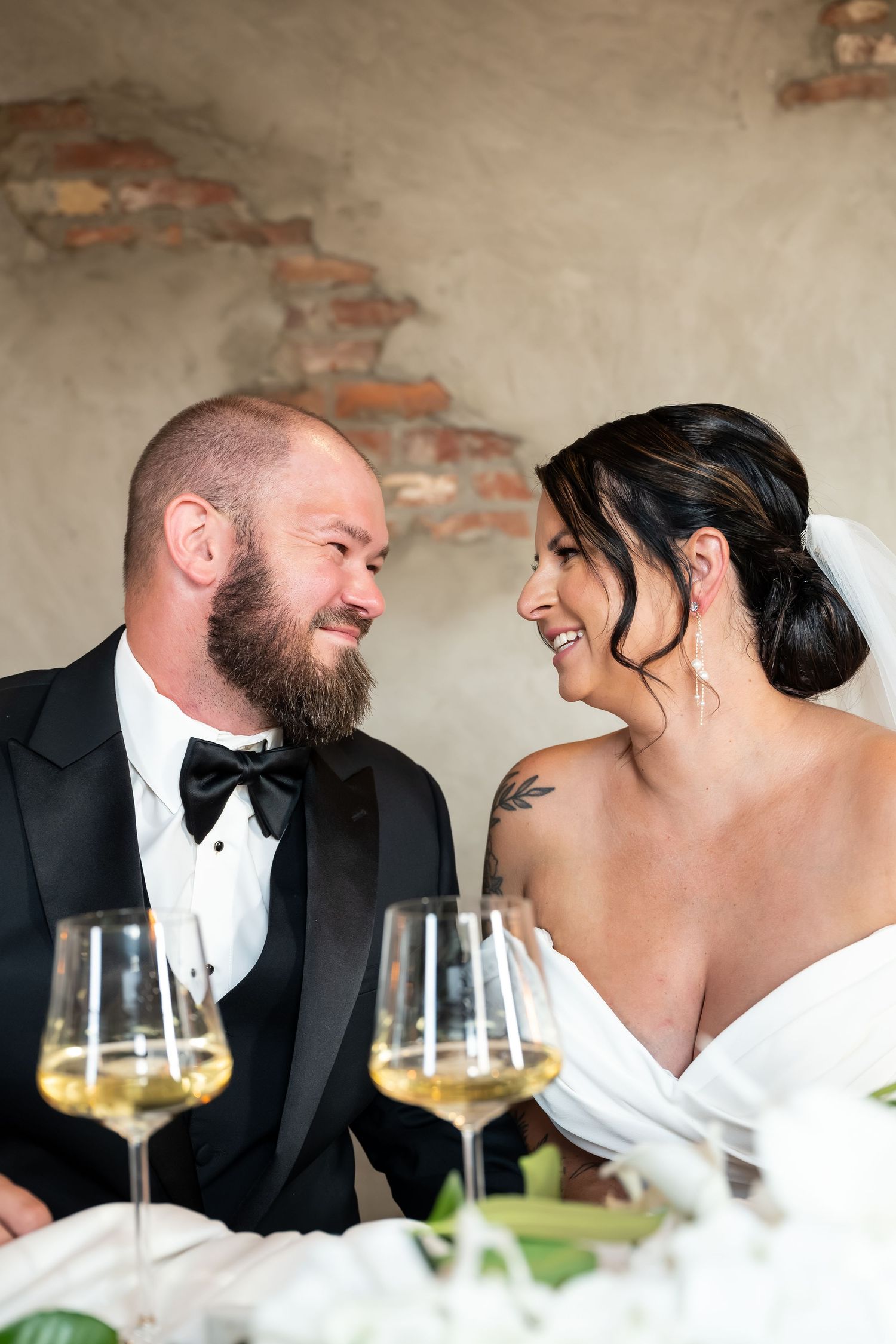A bride and groom share a loving glance at their wedding reception table with white wine glasses in the foreground.