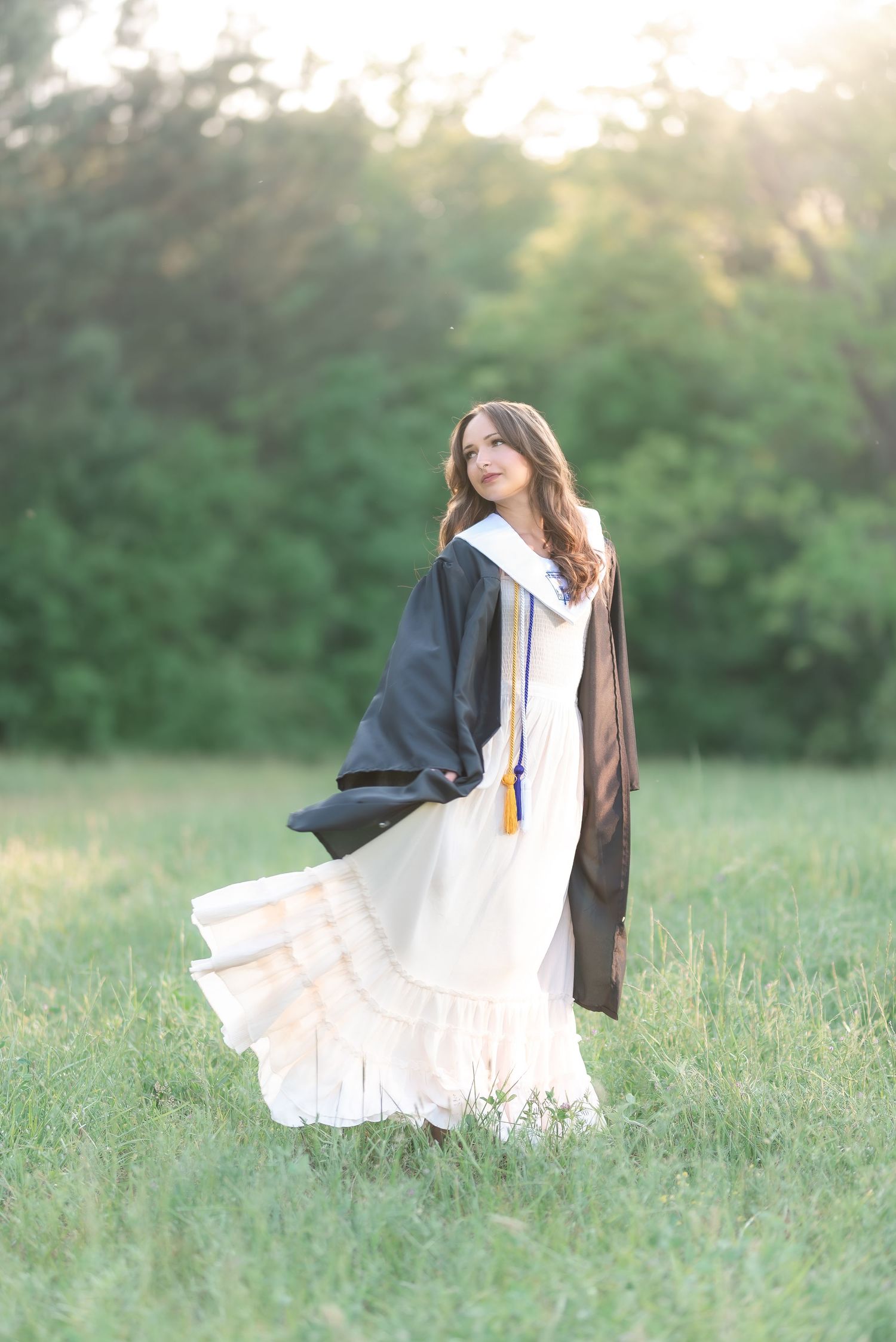Young woman in black graduation gown and cap over white dress standing in green meadow during golden hour.