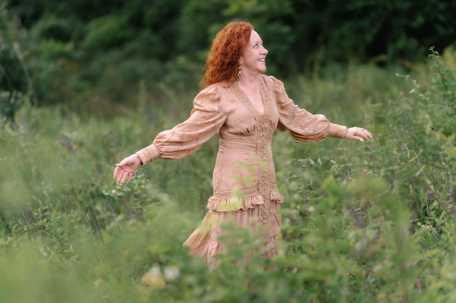 Red-haired woman in vintage beige dress twirling joyfully in a lush green meadow.