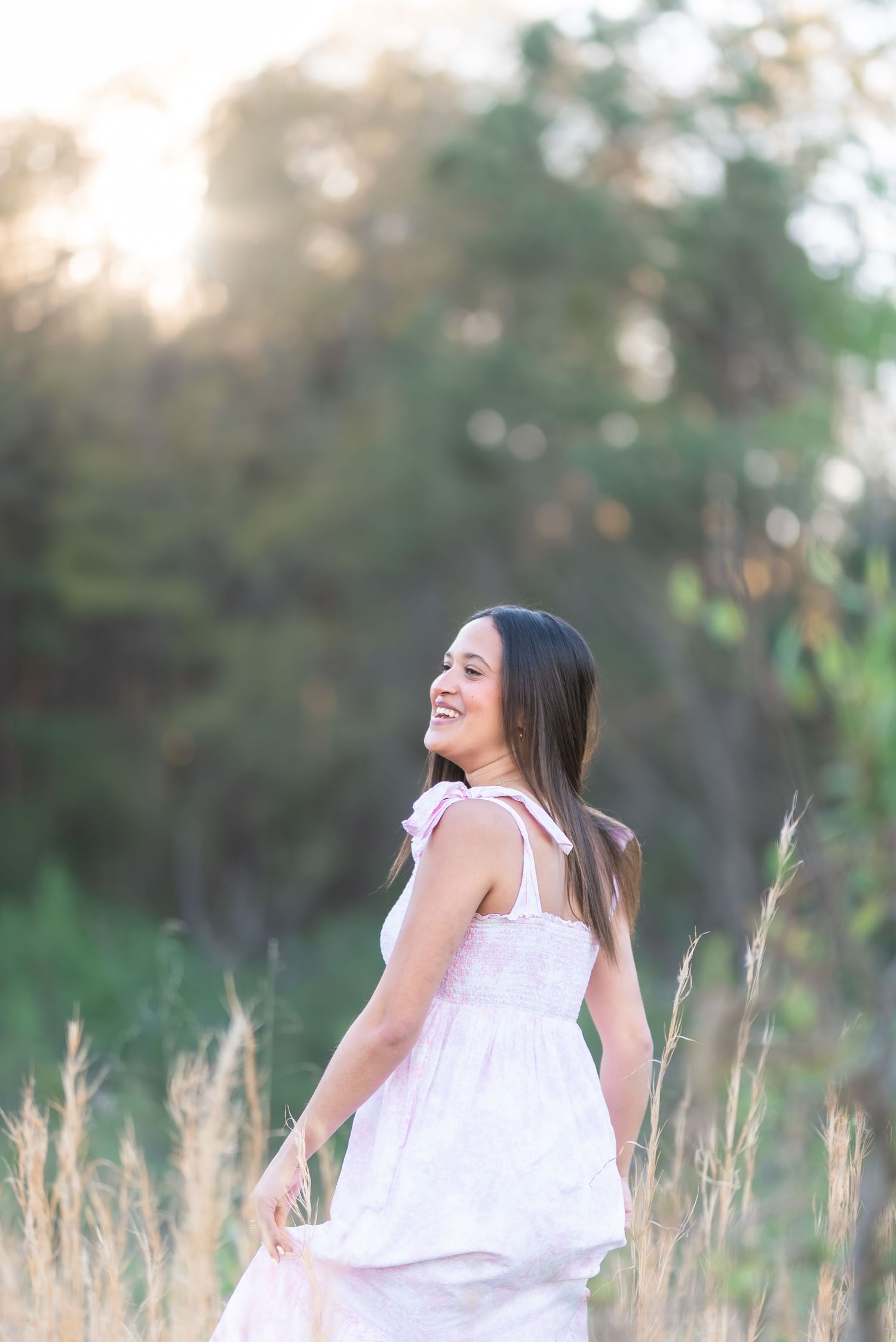 A smiling young woman in a white pink dress stands in a golden field at sunset with lush green trees behind her.