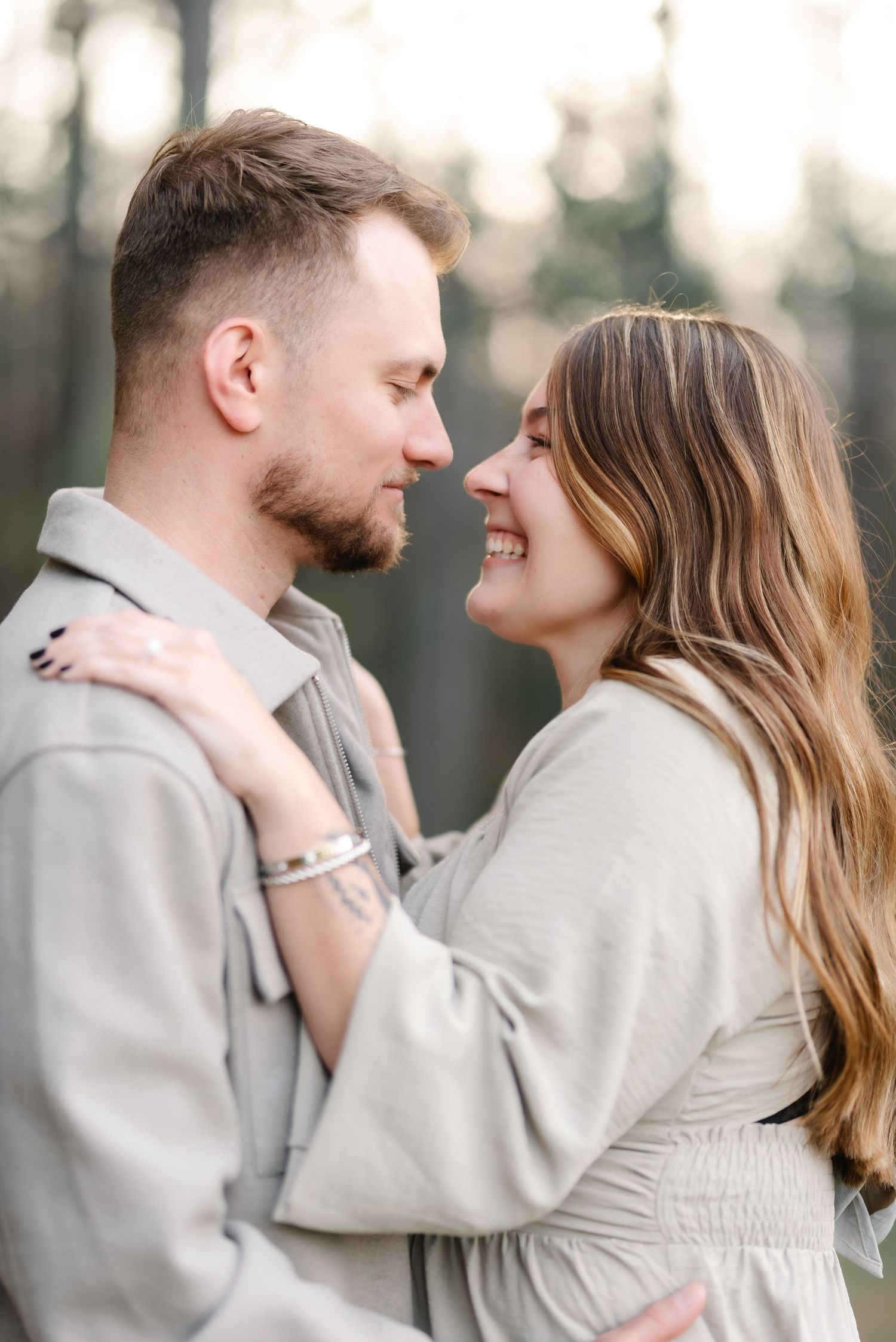 A smiling couple in neutral tones shares a tender moment outdoors, nearly touching foreheads in a romantic embrace.