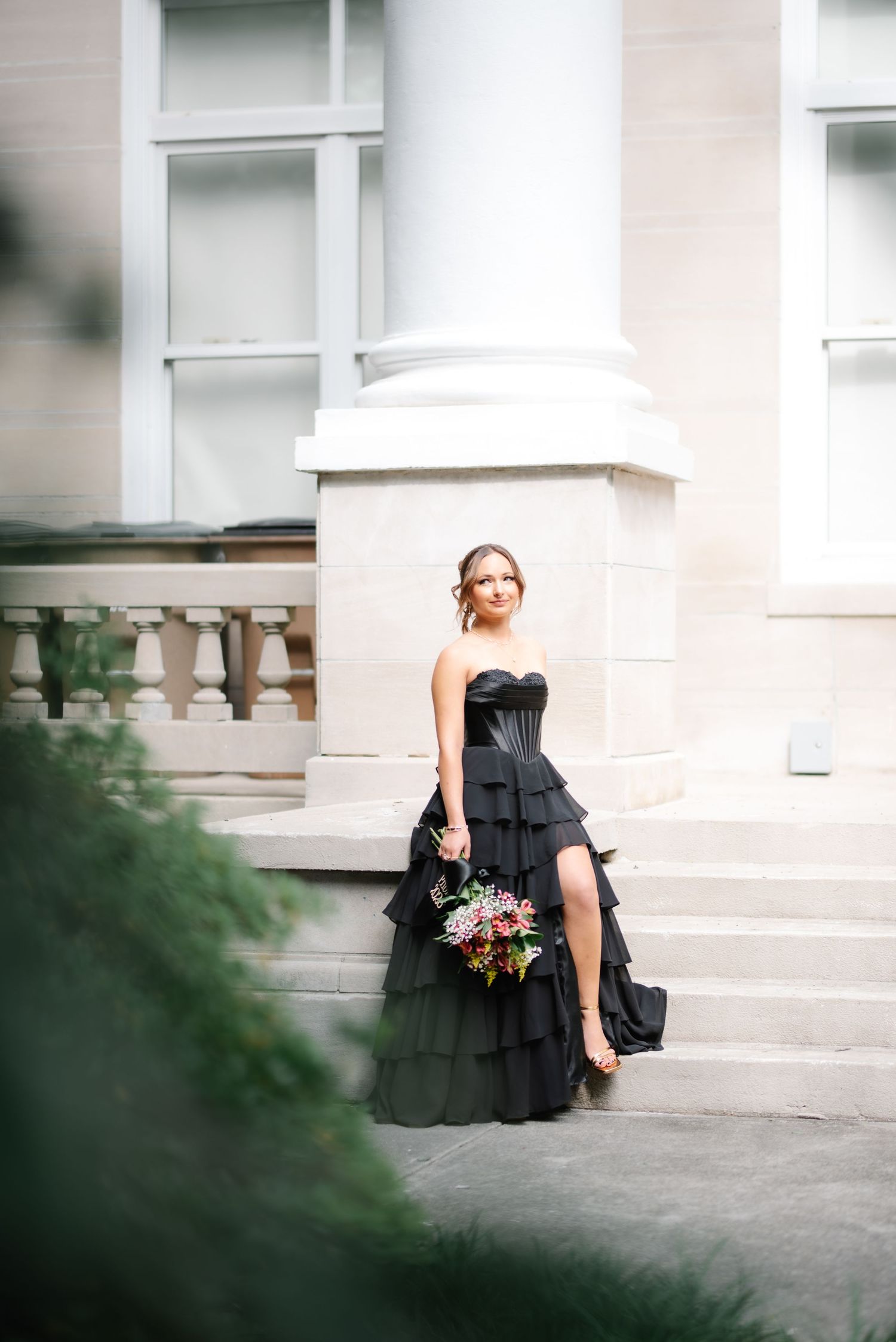 Woman in black strapless tiered gown holding floral bouquet on stone steps of elegant building.