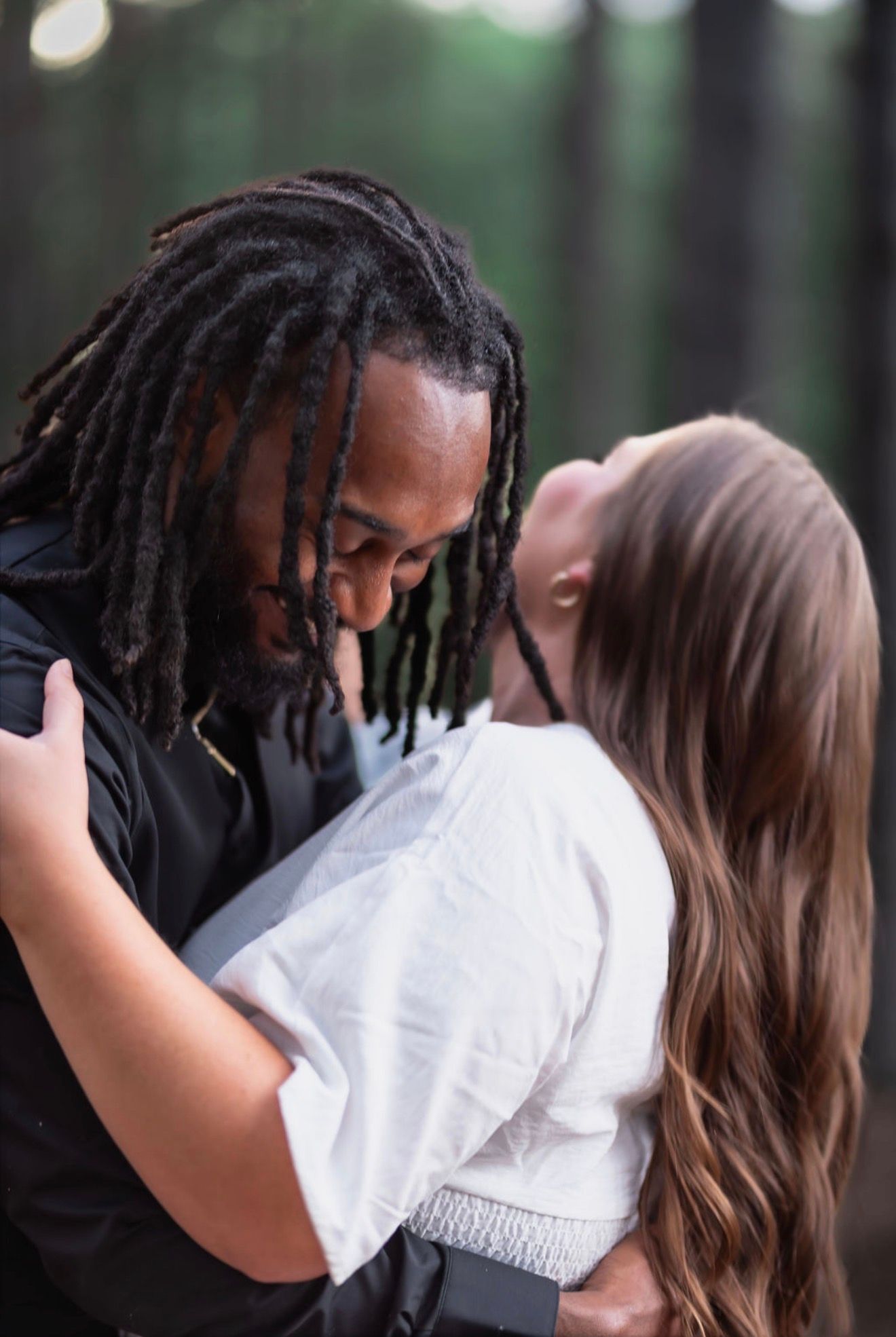 A couple shares a romantic moment in a forest, with the man's dreadlocks framing their faces as they lean in close.