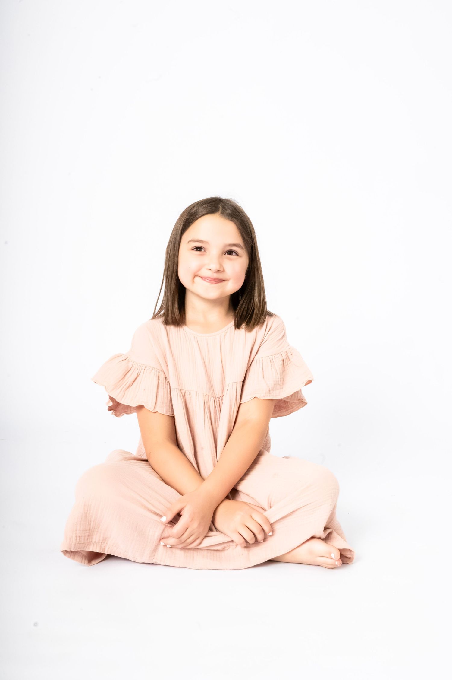 Young girl in pink ruffle dress sitting cross-legged on white background, smiling at camera.