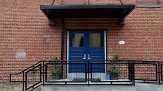 Brick building entrance with blue double doors, black metal railings, and potted plants under a dark canopy.