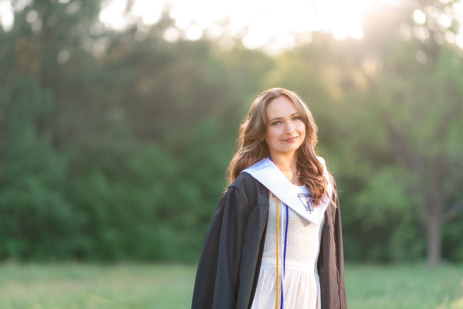 Smiling graduate in black cap and gown with white stole posing outdoors with green trees in background.