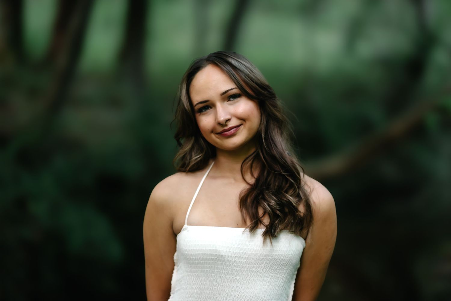 Young woman with long brown hair smiling outdoors, wearing a white halter top with lush green trees behind her.