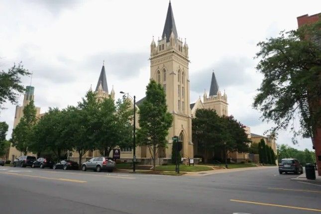 Historic Gothic church with tall spires surrounded by green trees on a city street corner on a cloudy day.